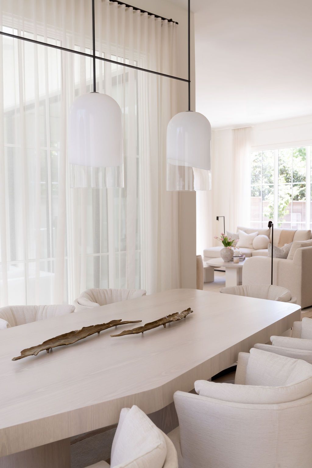 Minimalist white dining room with oval table, upholstered chairs, globe pendant lights, and sheer curtains.