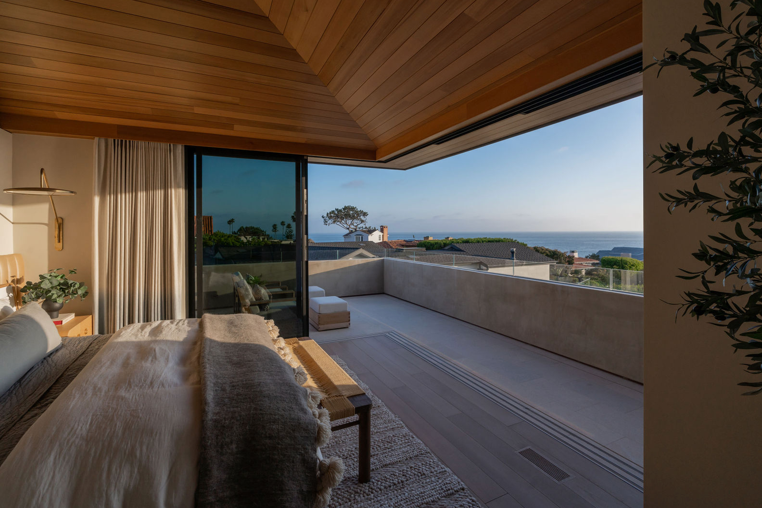 Master bedroom with wood ceiling, floor-to-ceiling glass doors, and panoramic ocean view at dusk.