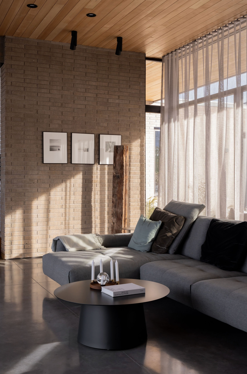 Modern living room with gray sectional sofa, round black coffee table, brick accent wall, and sheer floor-to-ceiling curtains.