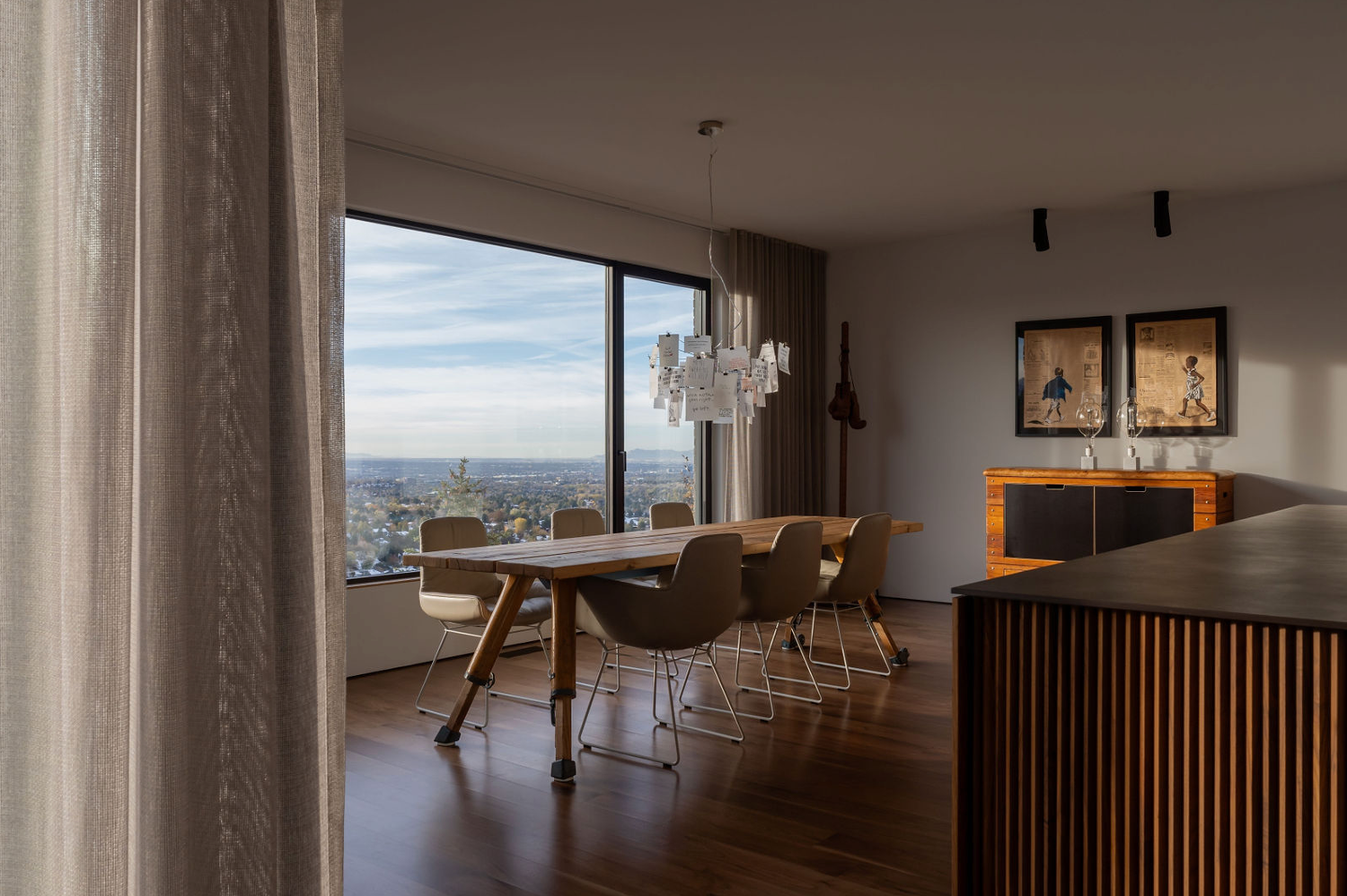 Elegant dining room with floor-to-ceiling windows offering city views, crystal chandelier, wooden table, and modern chairs.