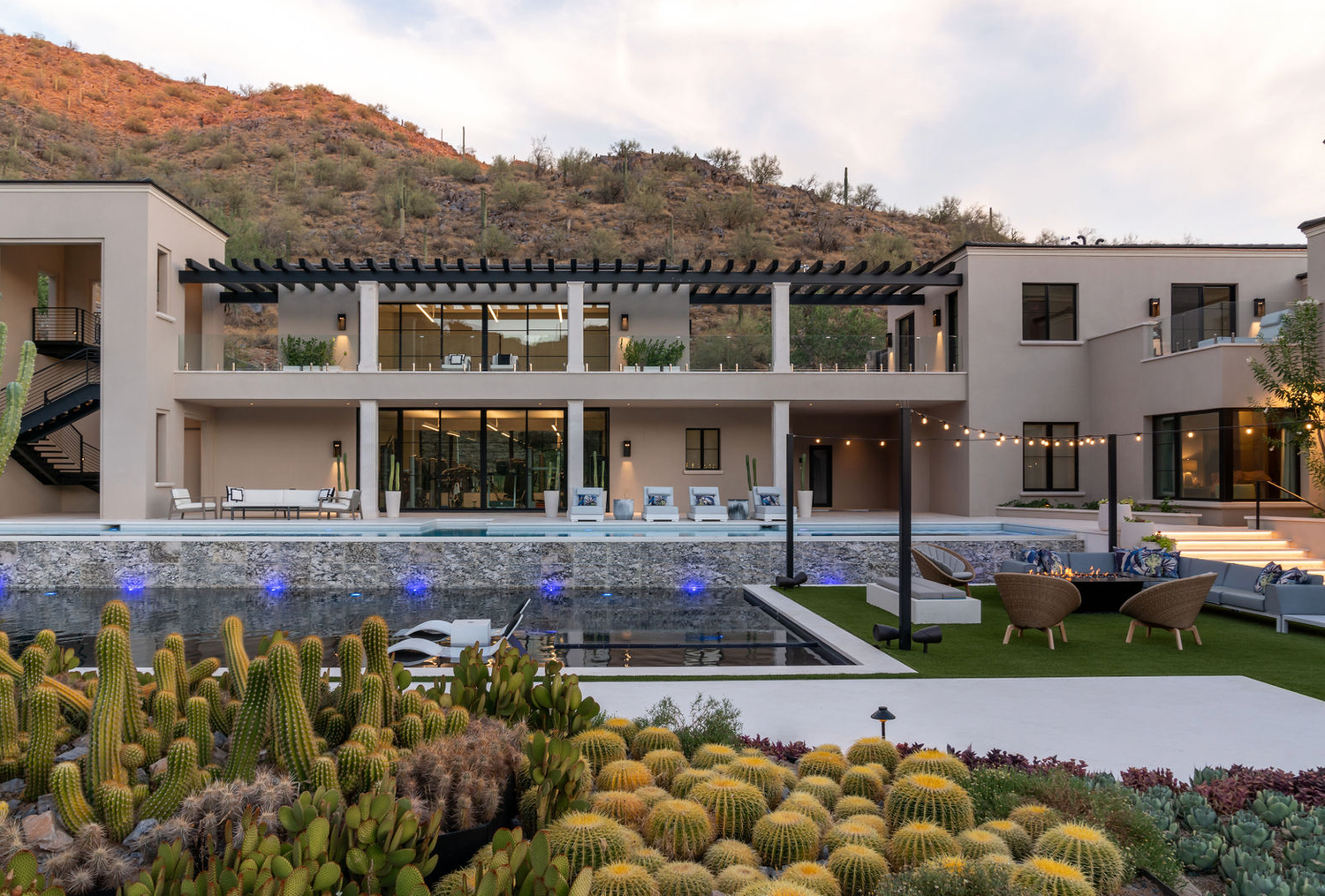 Modern desert estate with a pergola, infinity pool, cactus garden, and string lights at twilight.