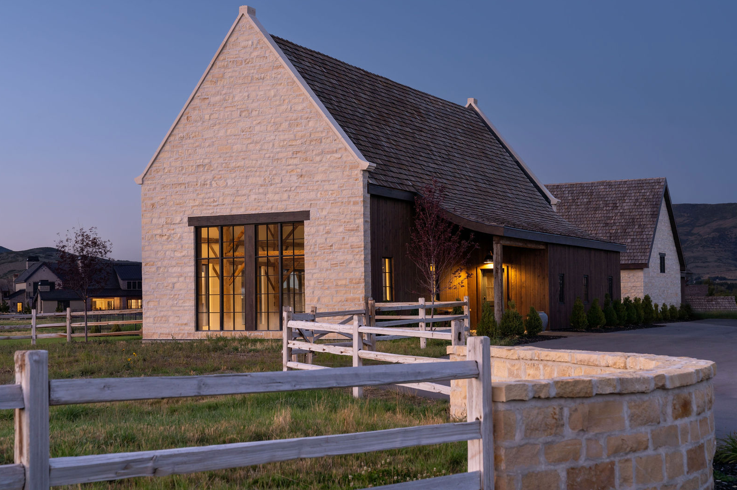 Elegant limestone barn with steep gabled roof and steel windows illuminated at dusk with white fence in foreground.
