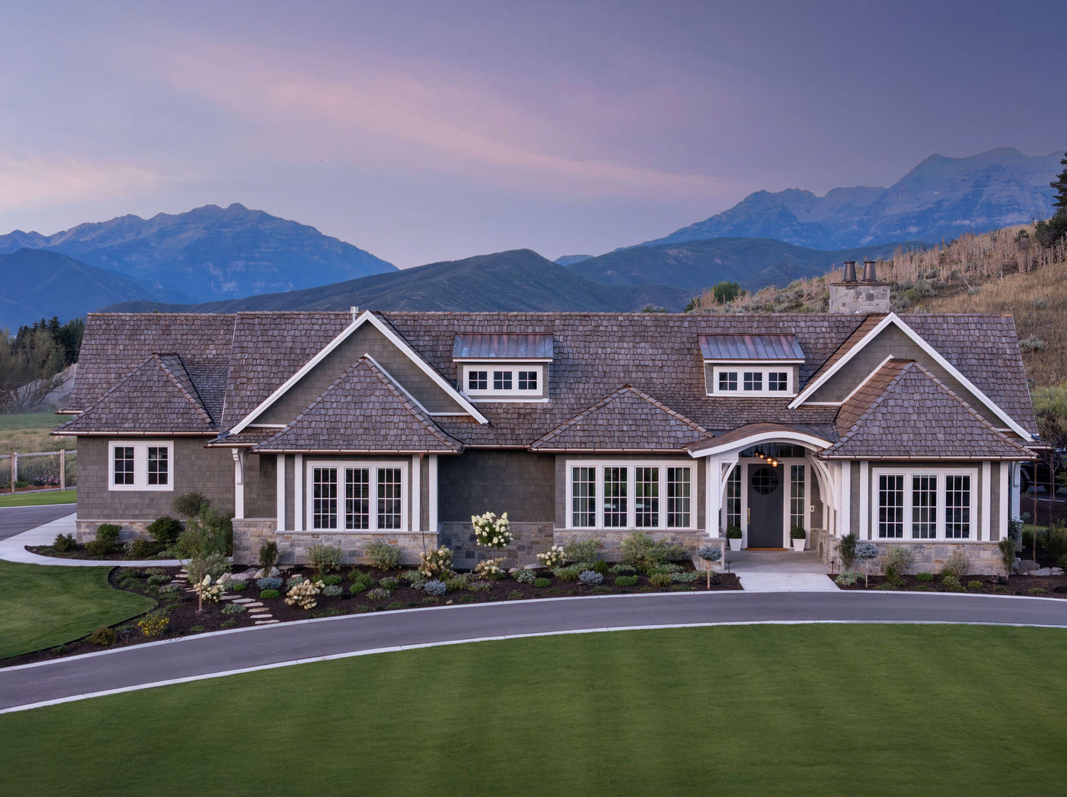 Grand craftsman ranch estate with cedar shake roof, dormer windows, and stunning Rocky Mountain backdrop at dusk.