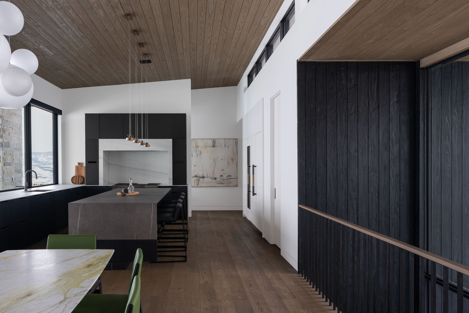 Sleek modern kitchen with black cabinetry, wood ceiling, dark island, and green dining chairs on hardwood floors.