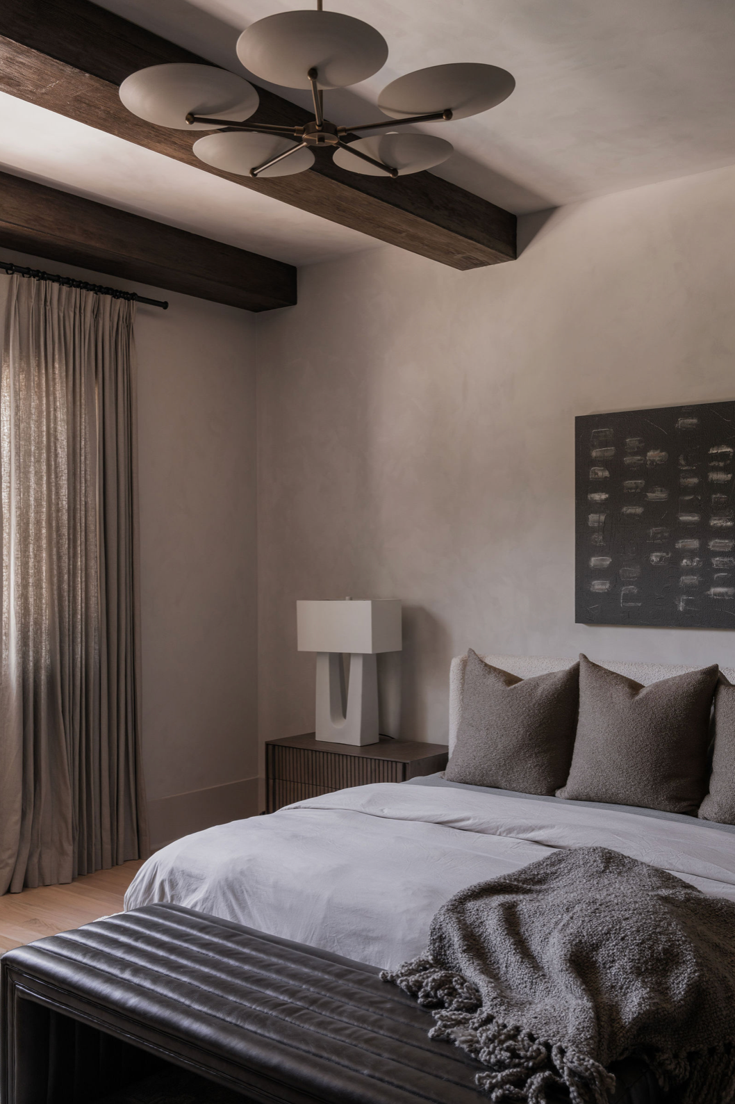 Moody bedroom with plaster walls, dark wood beams, sculptural ceiling light, and layered gray bedding.