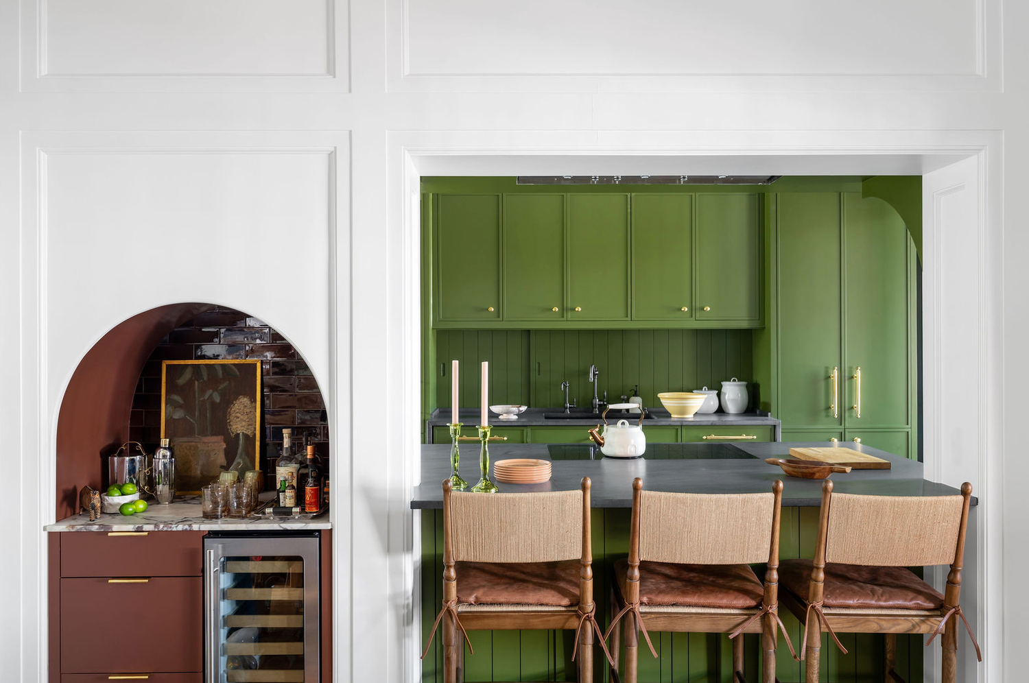 Bold green kitchen with arched bar nook, dark countertops, rattan barstools, and brass hardware.