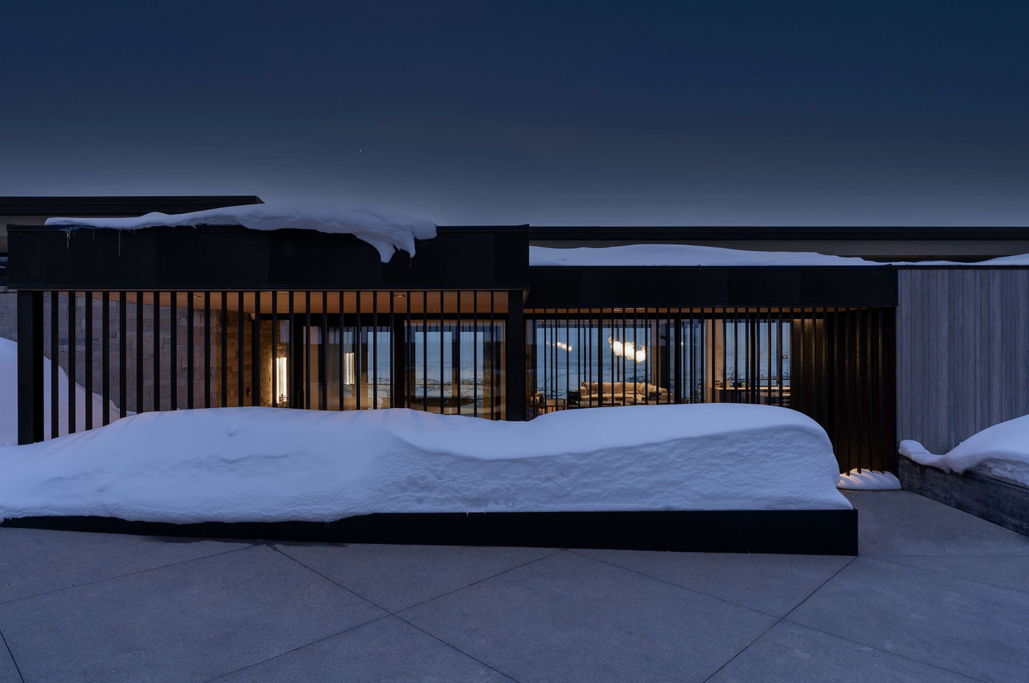 Contemporary home exterior with dark vertical slats and snow-covered patio photographed at night.