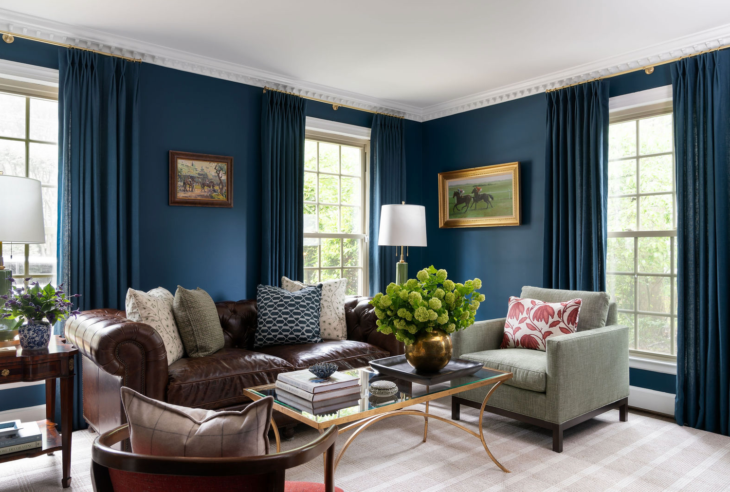 Sophisticated navy blue living room with leather sofa, gray armchairs, gold coffee table, and tall draped windows.
