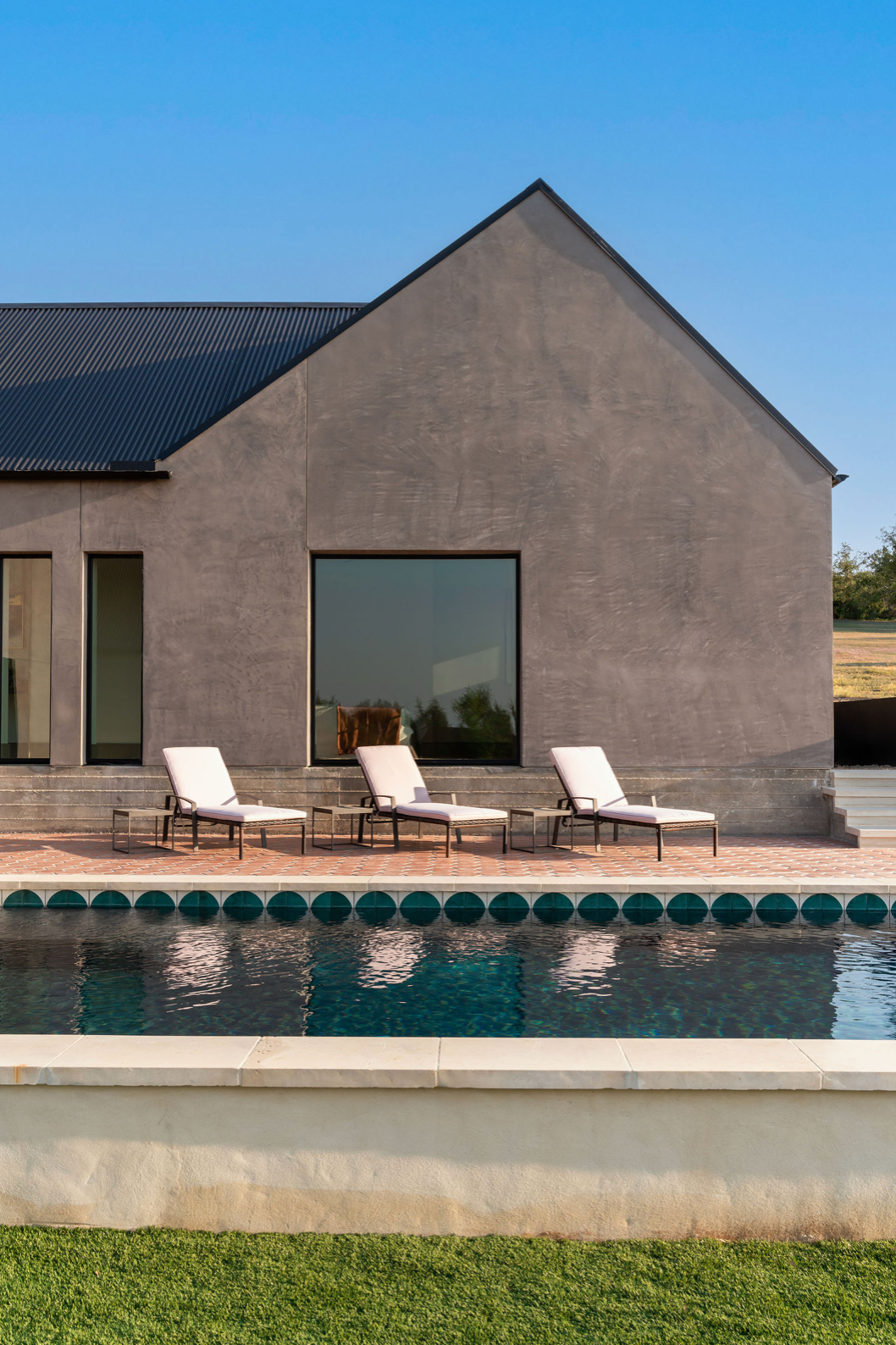 Minimalist stucco gable home with dark metal roof, poolside lounge chairs, and teal-tiled pool edge on sunny day.