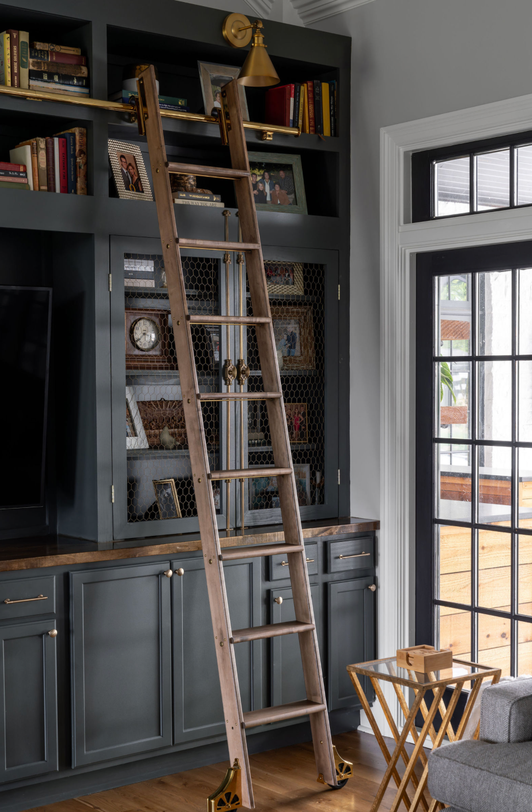 Dark gray built-in bookcase with library ladder, glass cabinet doors, and brass hardware in elegant home office.