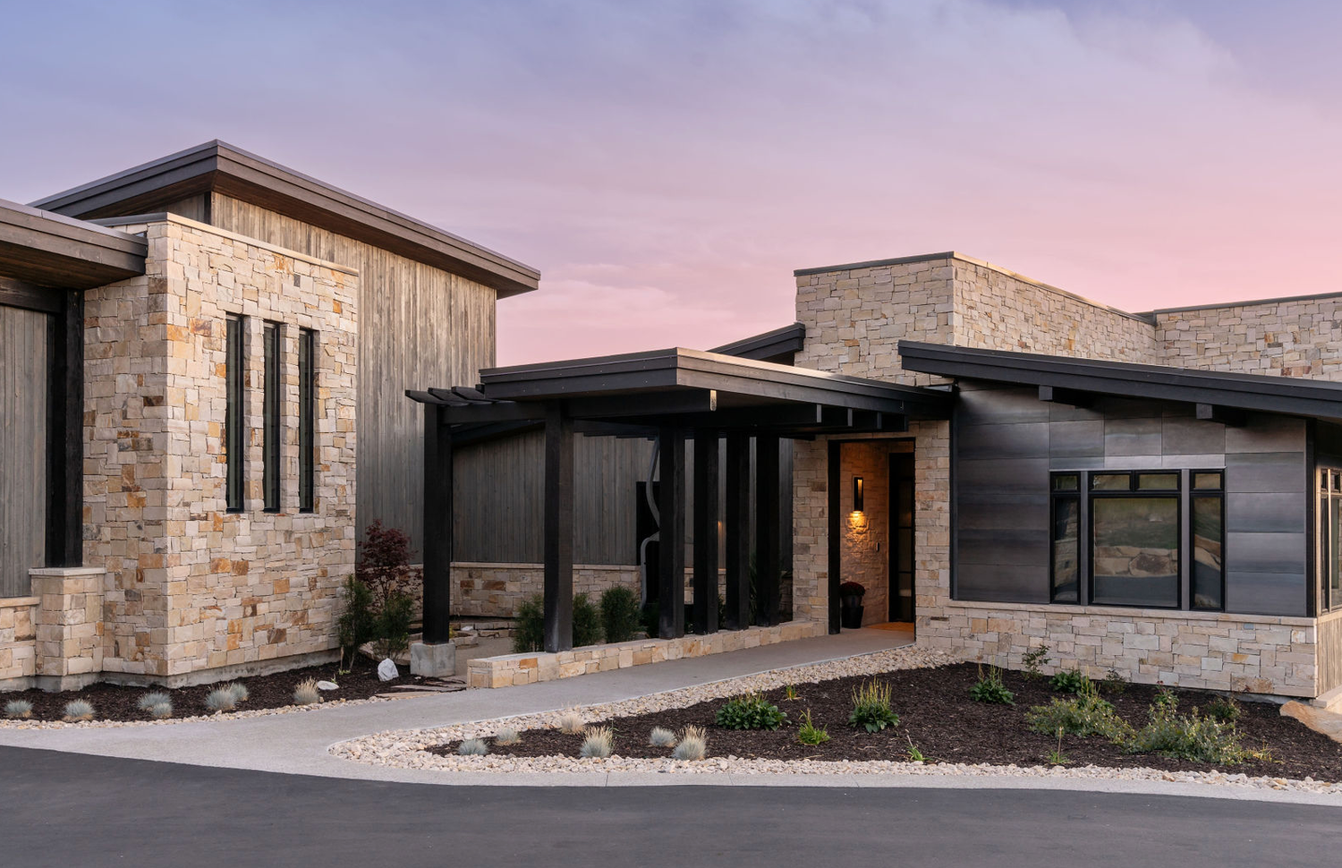 Contemporary ranch-style home combining natural stone, dark wood siding, and black metal roof at twilight.