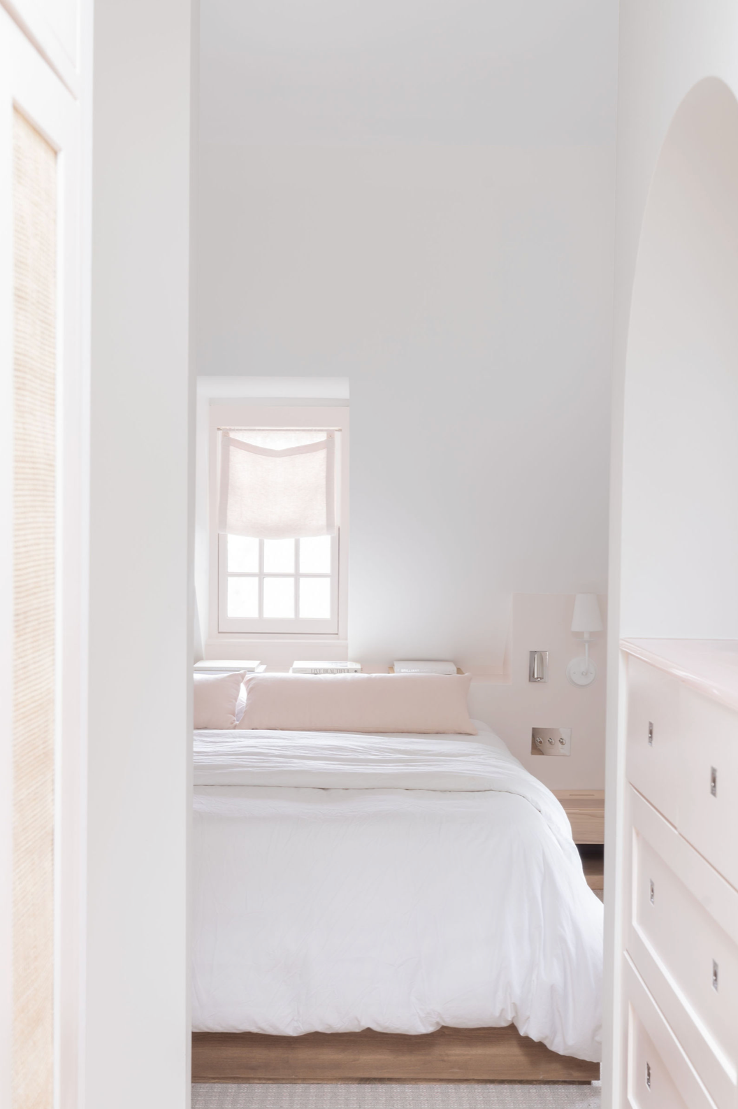 Bright minimalist bedroom viewed through archway with white bedding, blush accents, and natural wood dresser.