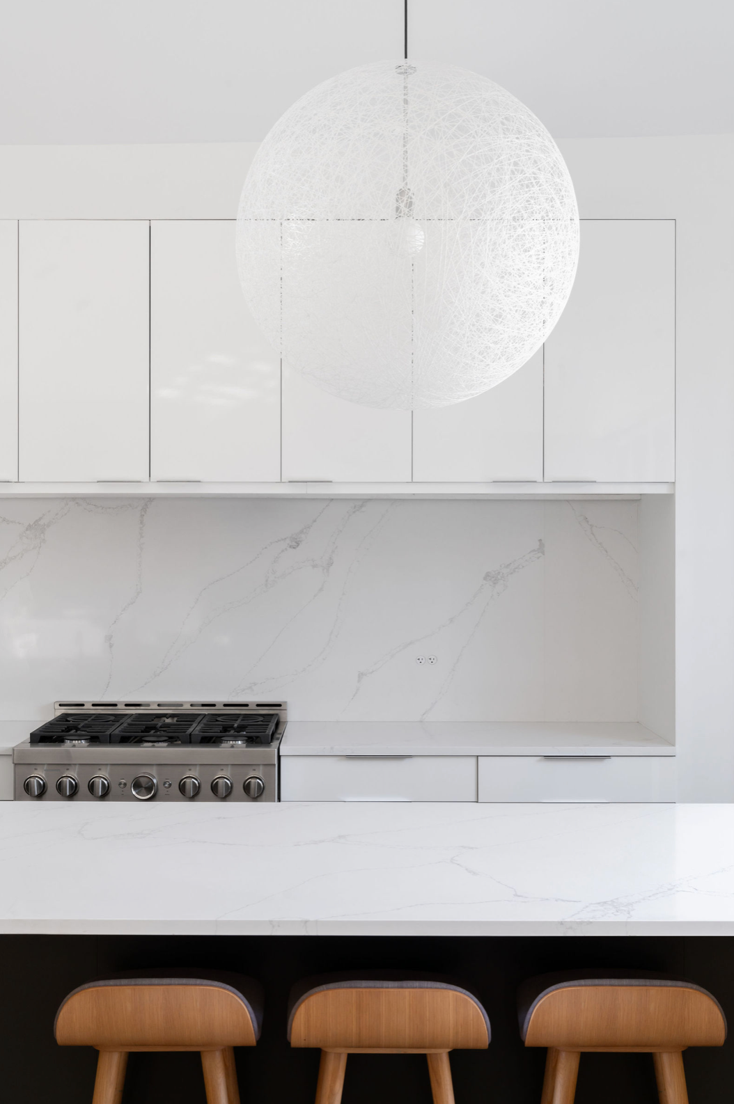 Minimalist white kitchen with marble backsplash, globe pendant light, gas range, and warm wood bar stools.