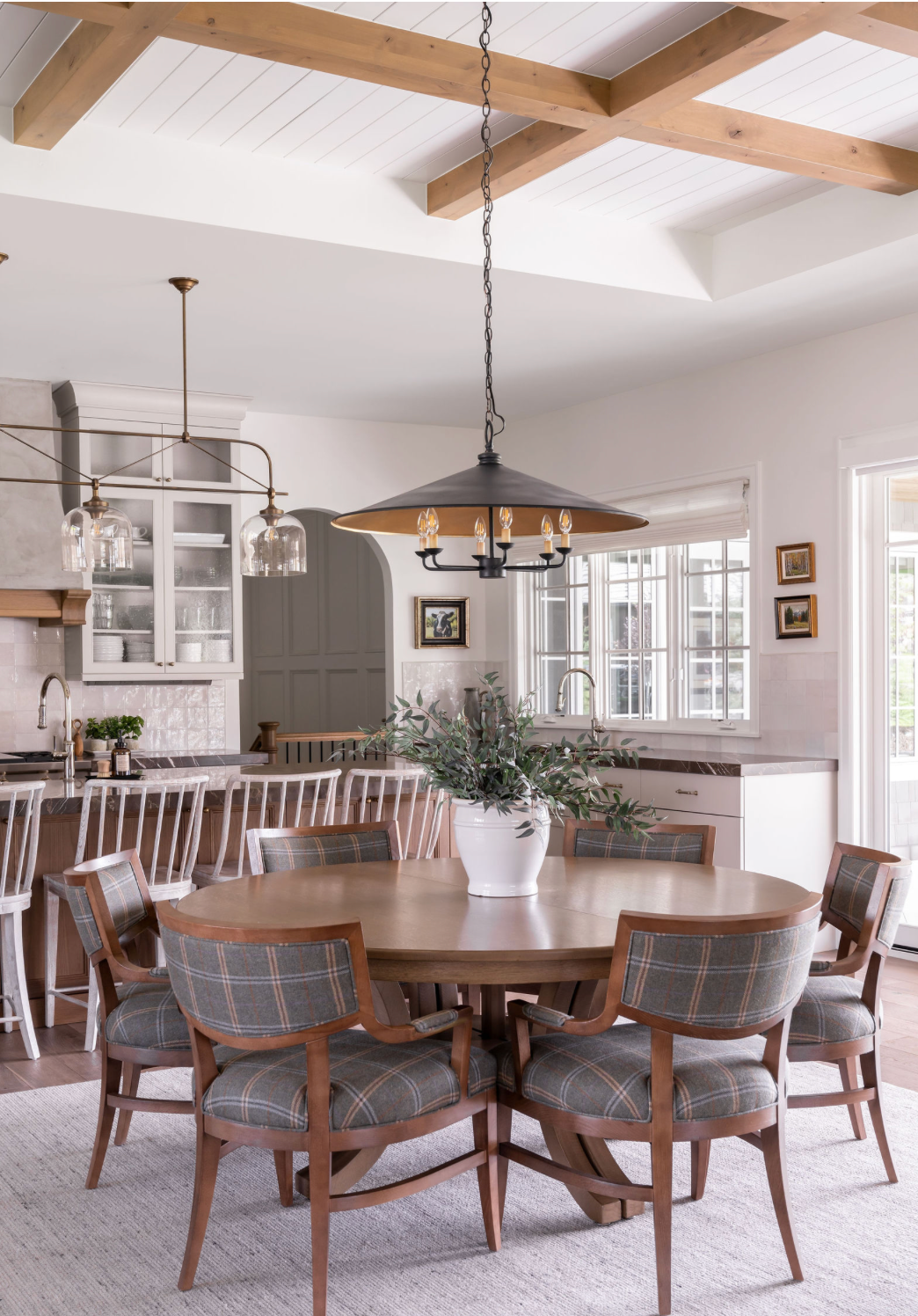 Farmhouse dining room with round wood table, plaid upholstered chairs, coffered ceiling, and black pendant light.