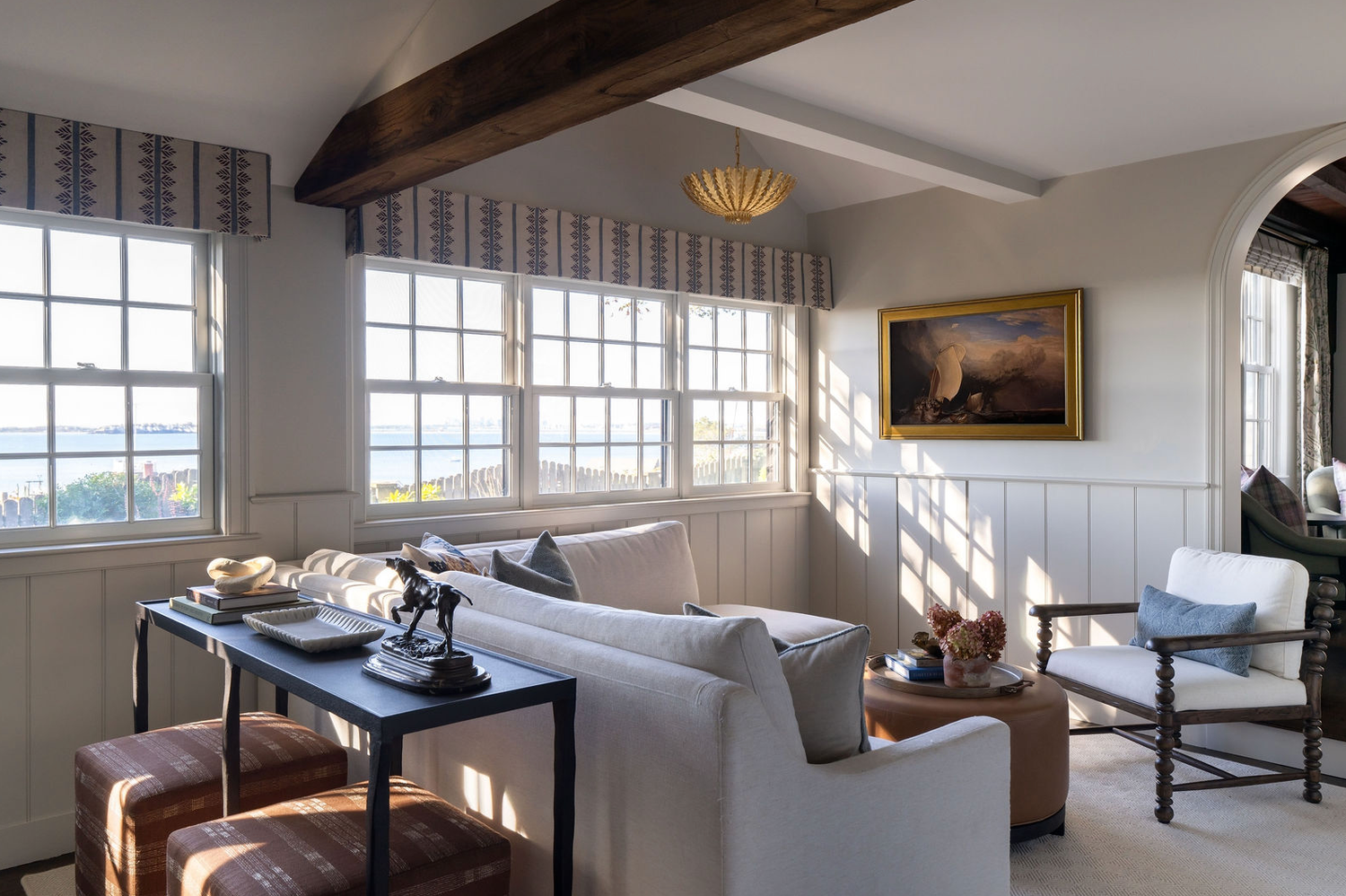 Cozy coastal living room with exposed beam, large windows, neutral sofa, and classic oil painting.