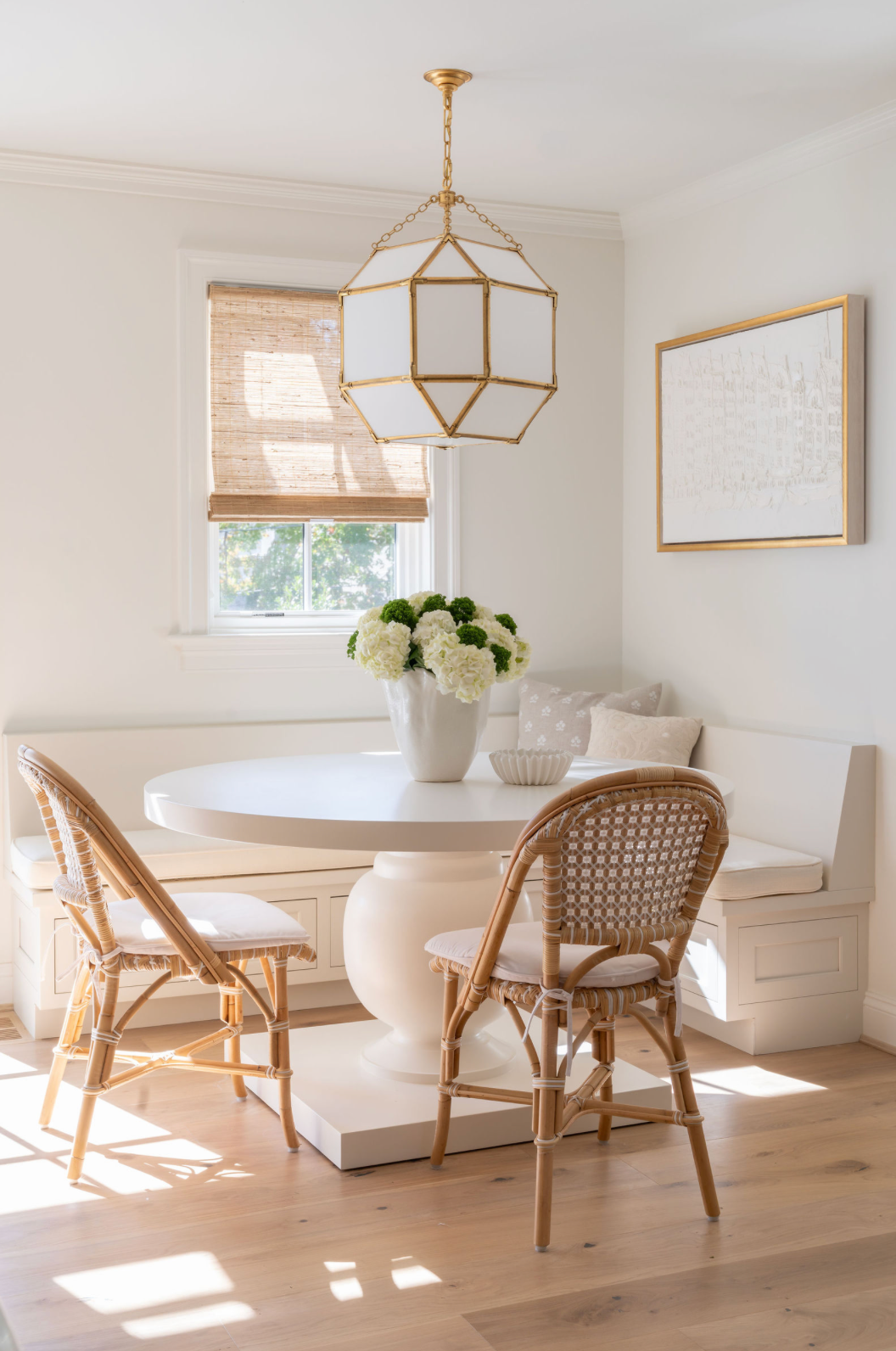 Elegant breakfast nook with round white pedestal table, rattan chairs, and gold geometric pendant light.
