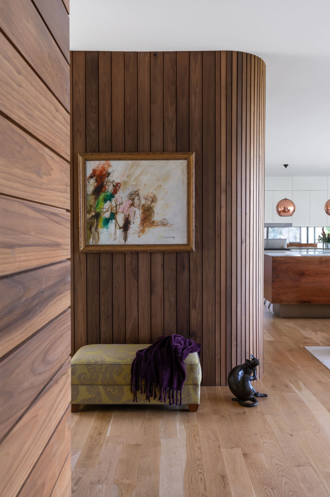 Curved walnut wood slat wall with abstract painting above a green velvet ottoman with purple throw in modern entryway.
