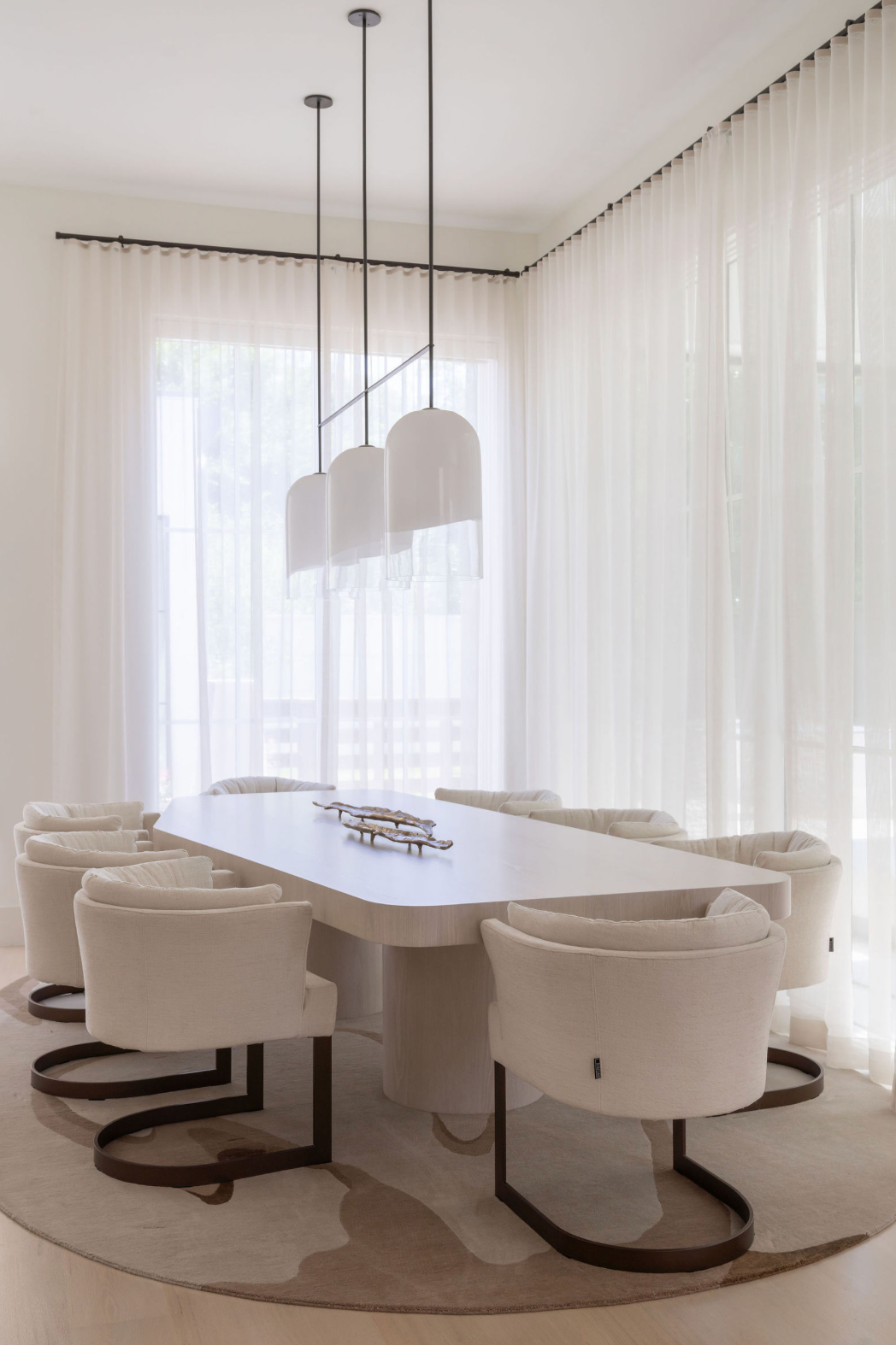 Elegant all-white dining room with upholstered chairs, pedestal table, pendant lights, sheer curtains.
