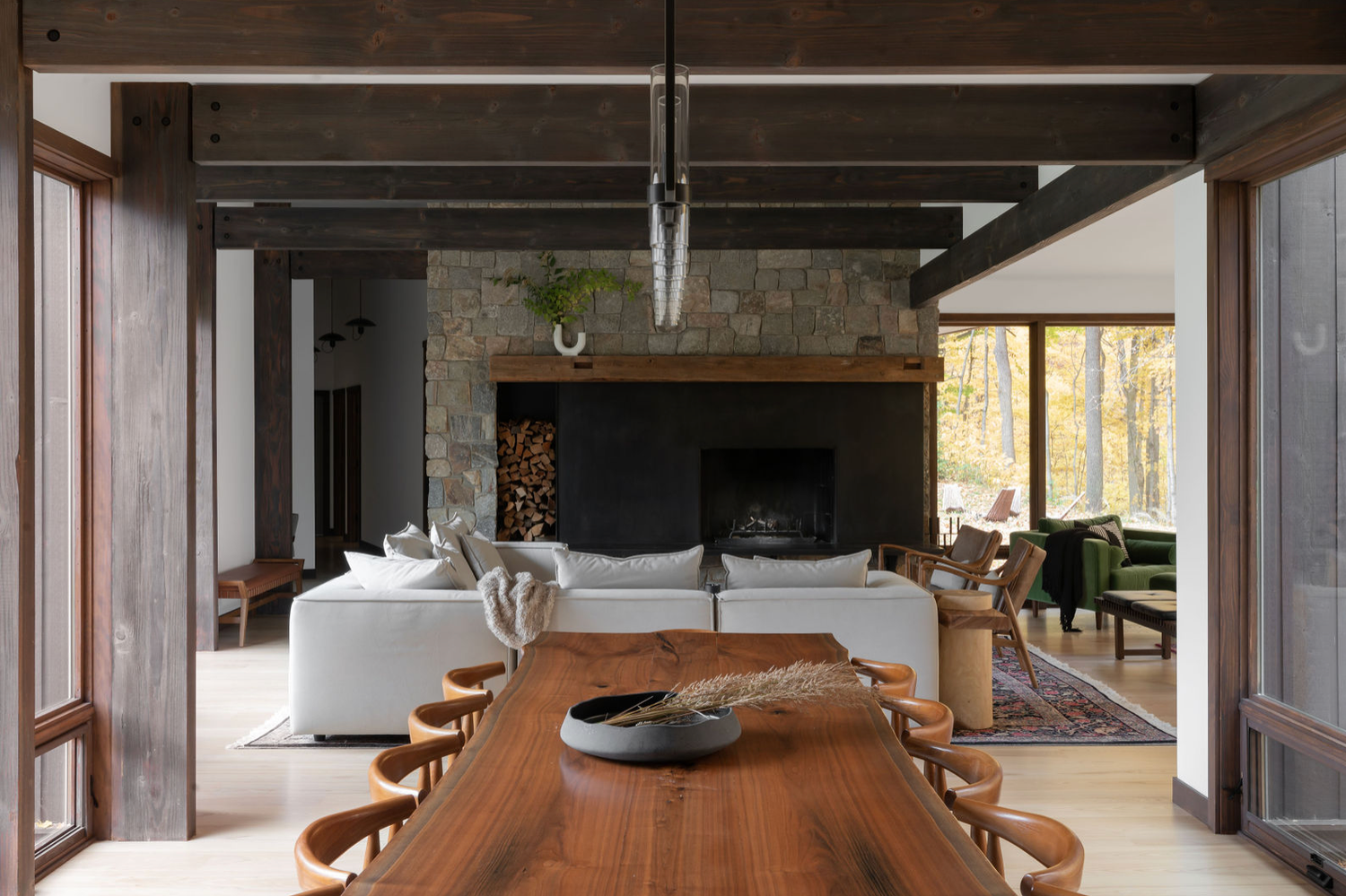 Rustic open-plan living and dining room with a live-edge wood table, stone fireplace, white sofas, and forest views.