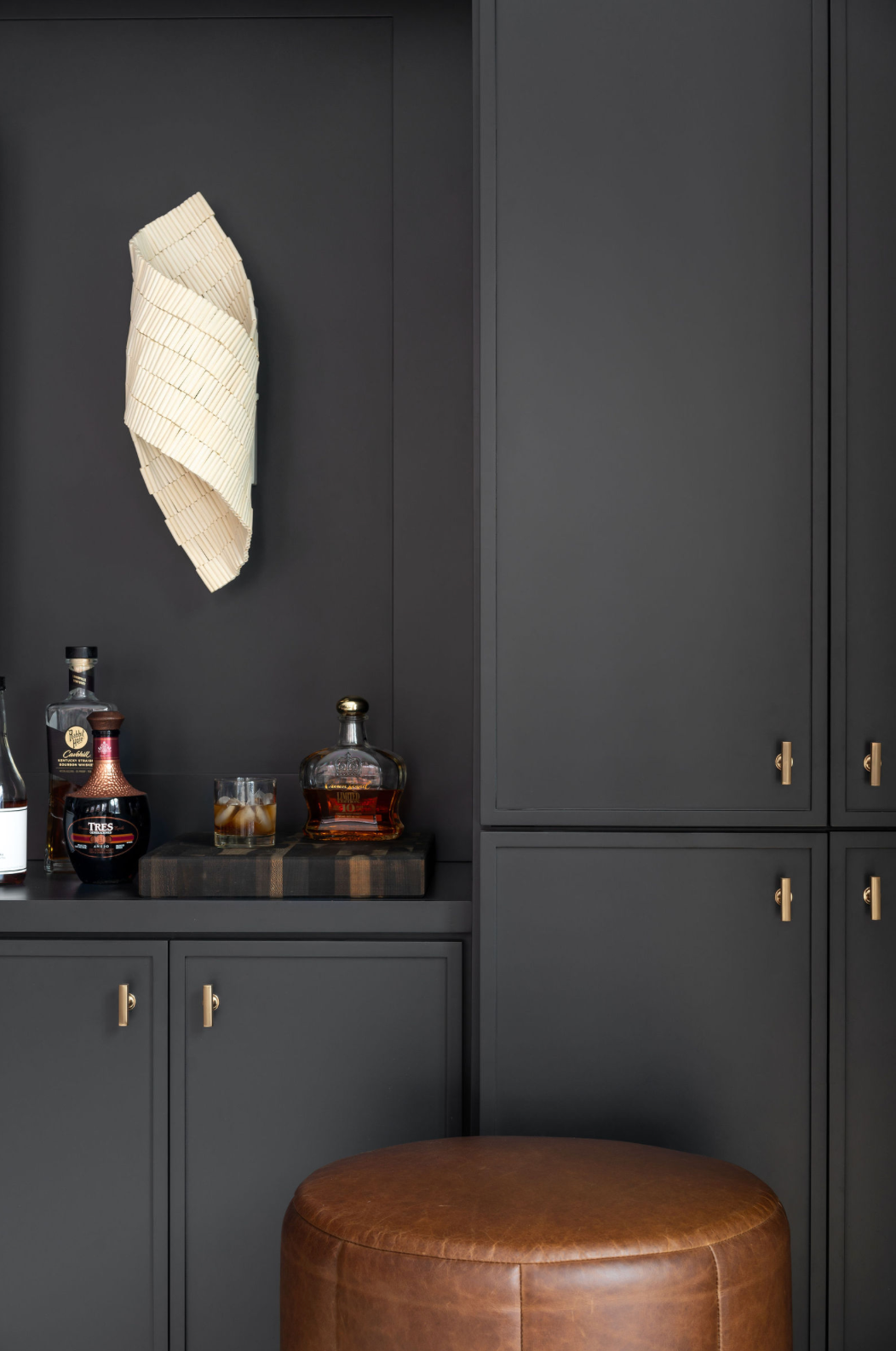 Dark charcoal built-in bar cabinet with brass hardware, liquor bottles, and leather ottoman.