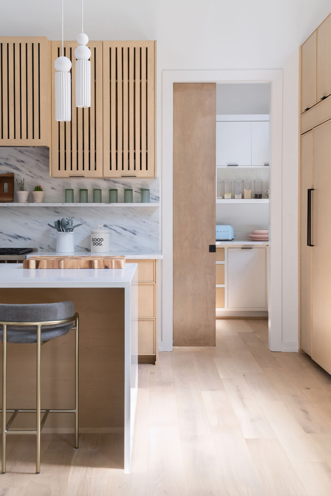 Modern kitchen with light wood slatted cabinets, marble backsplash, island with barstool, and globe pendant lights.