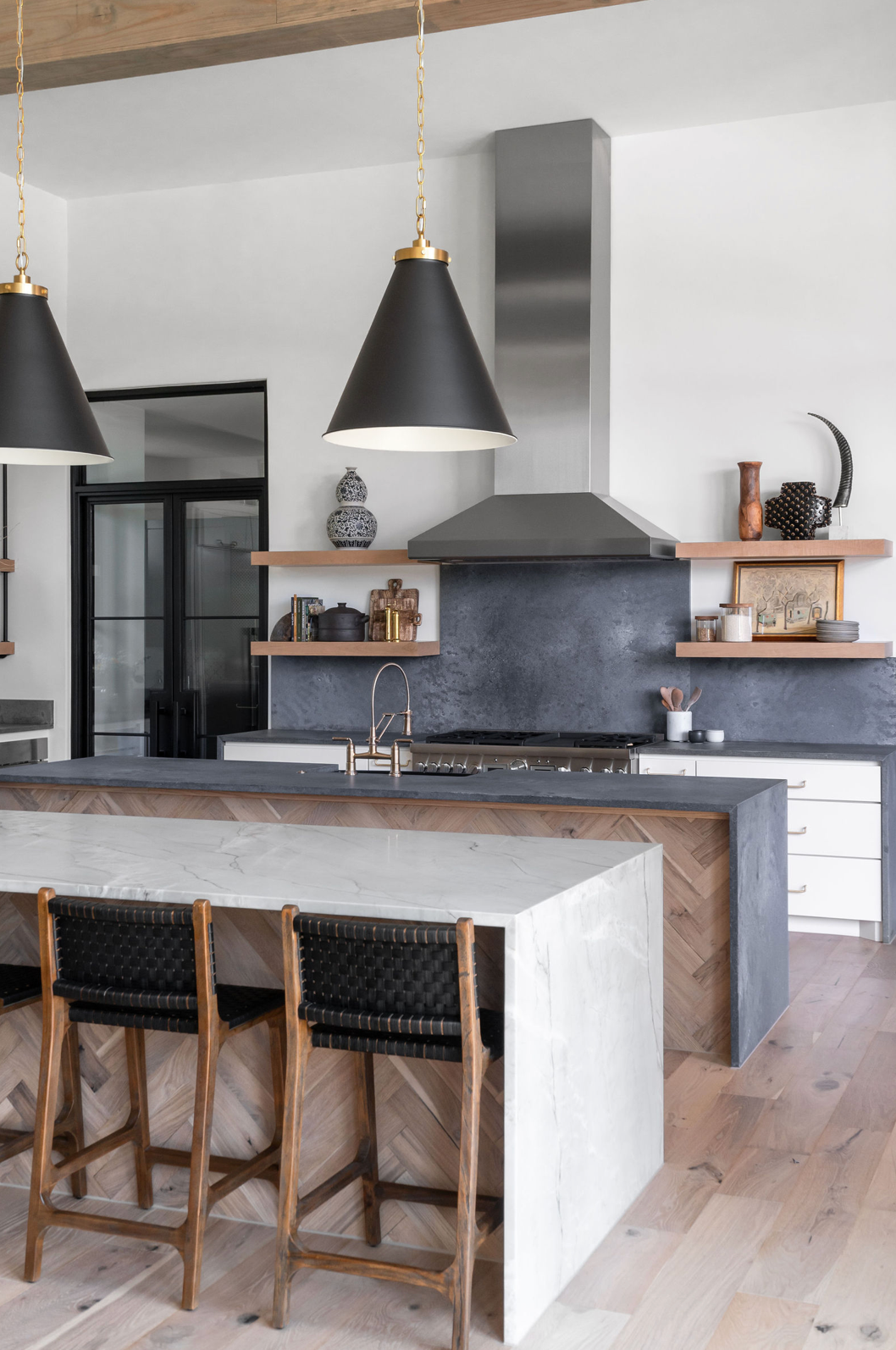 Modern kitchen with black cone pendant lights, concrete backsplash, herringbone wood island base, and open wood shelving.
