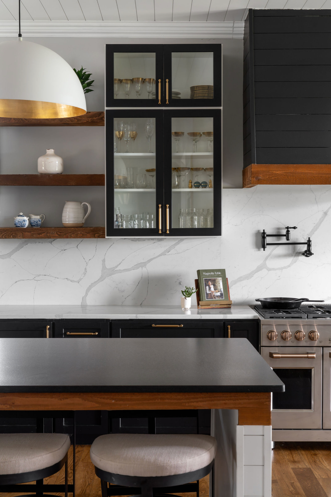 Black kitchen cabinets with glass doors, marble backsplash, open wood shelves, and gold dome pendant light.