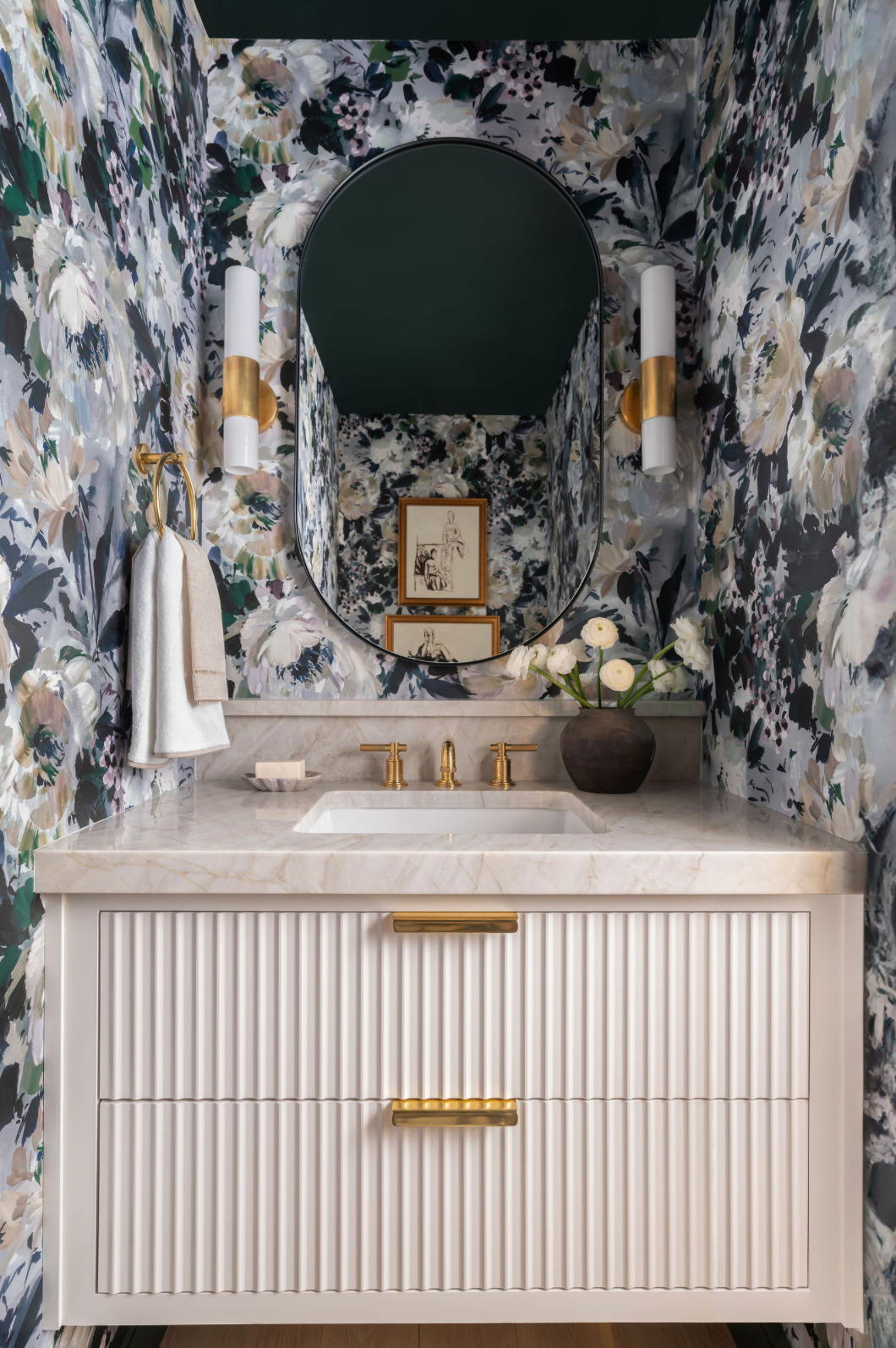 Bold floral wallpaper powder room with fluted white vanity, round mirror, and gold fixtures.