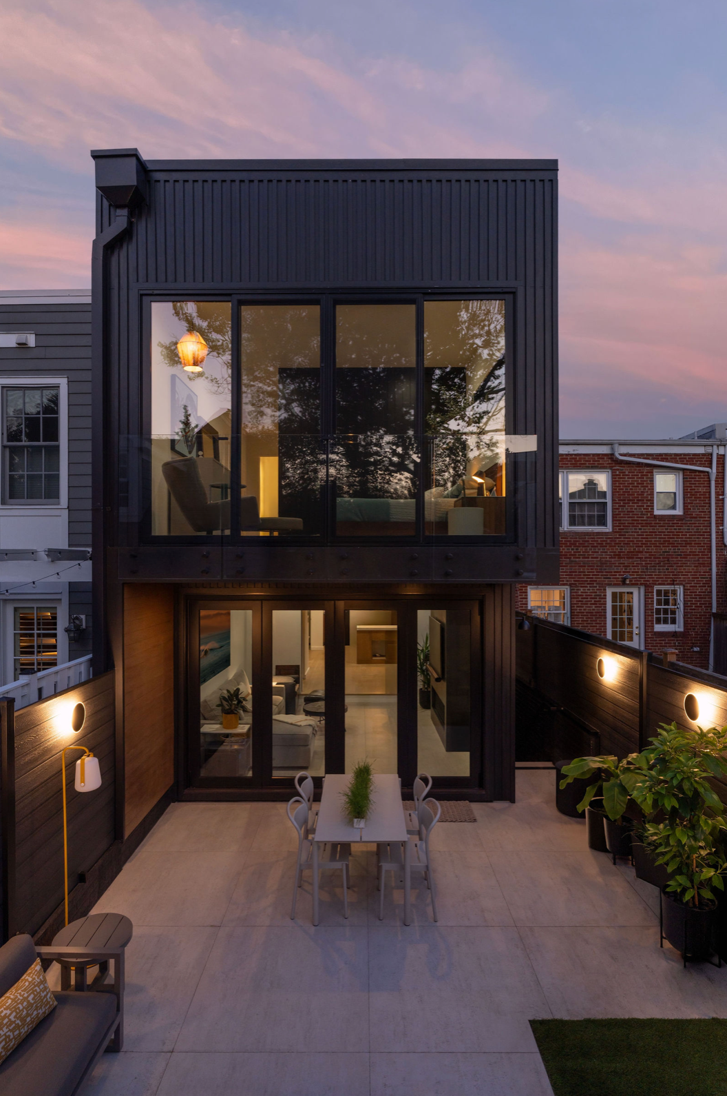 Modern black townhouse exterior with floor-to-ceiling windows and patio dining area photographed at sunset.
