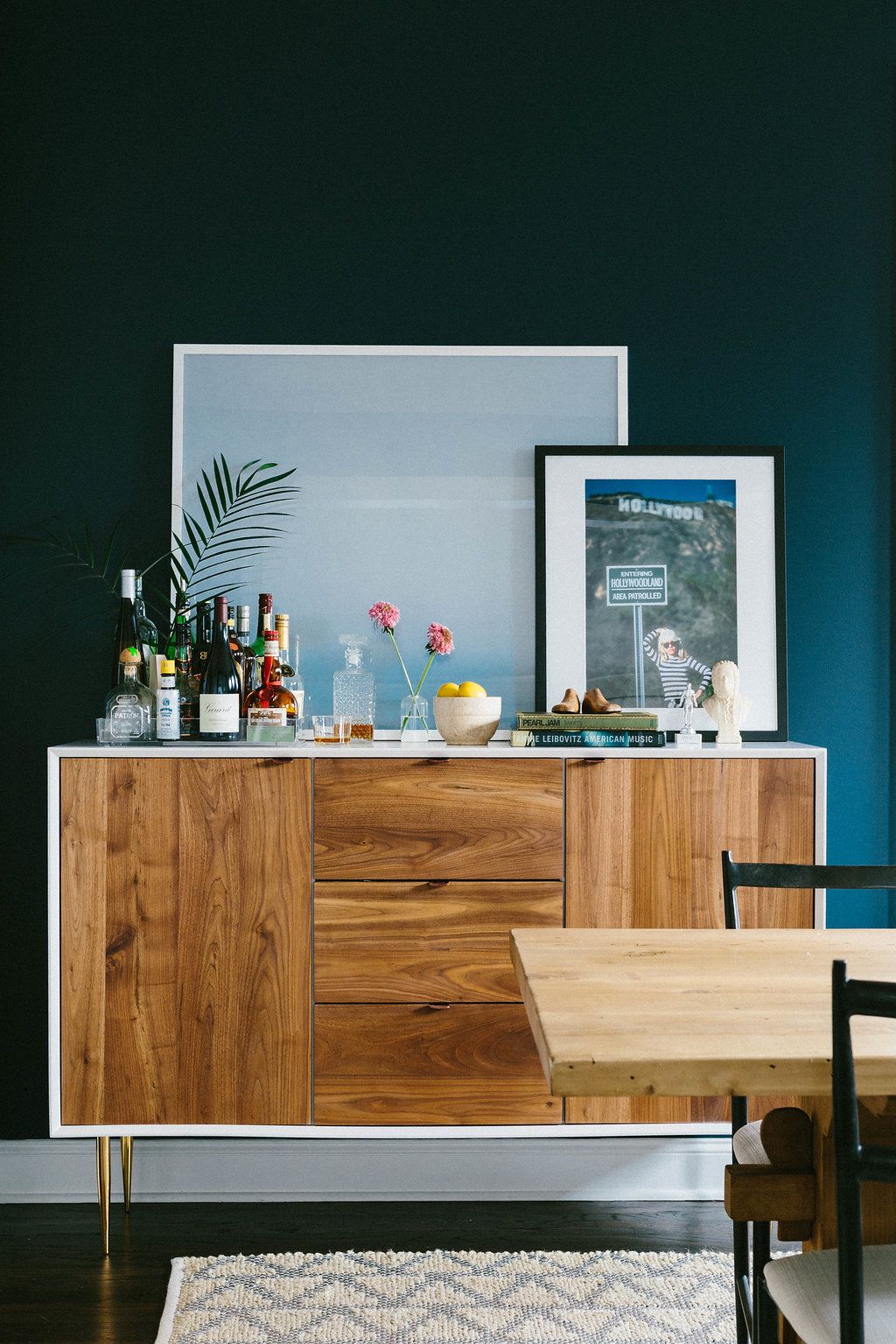 Dark teal dining room with walnut sideboard styled as a bar cart, framed art prints, and wood dining table.