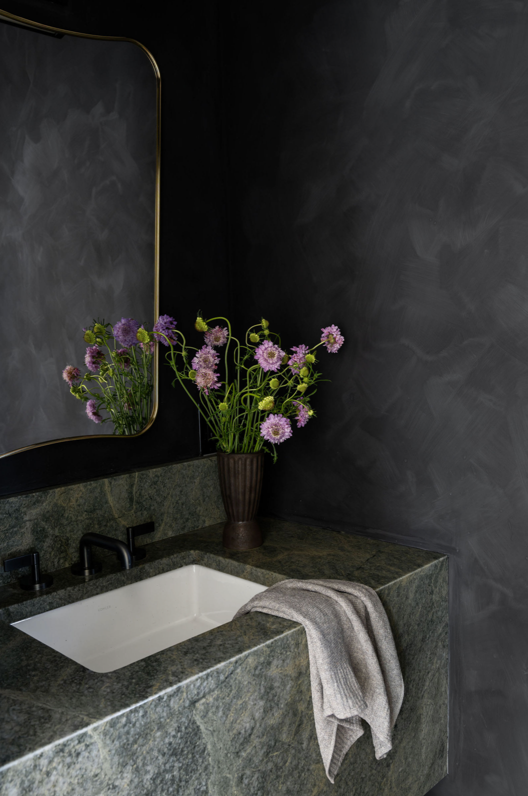 Dark moody powder room with green marble sink, black walls, gold mirror, and purple flowers.
