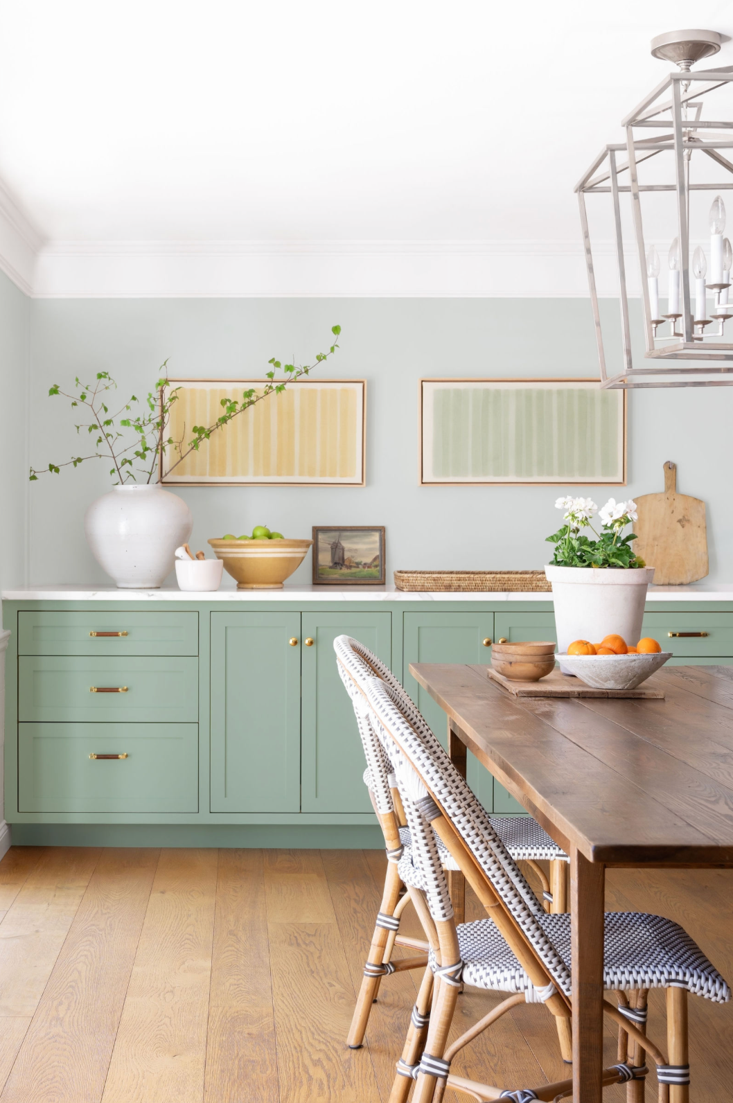 Sage green kitchen cabinets with brass hardware, white countertops, bistro chairs, and wood dining table.