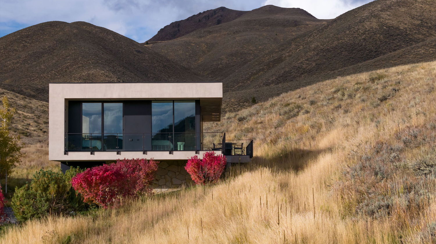 Minimalist modern home nestled in hillside landscape with red shrubs, golden grasses, and dramatic mountain backdrop.