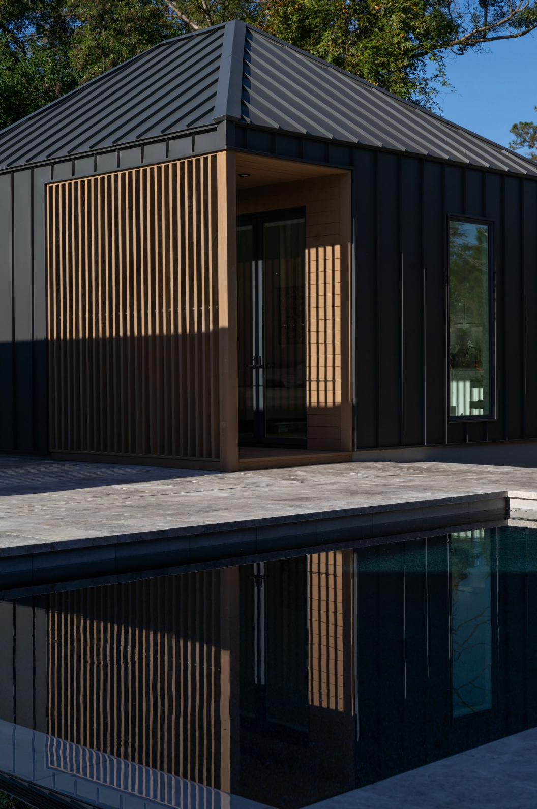 Sleek black pool house with vertical wood slat accents, metal roof, and dark pool reflection at midday.