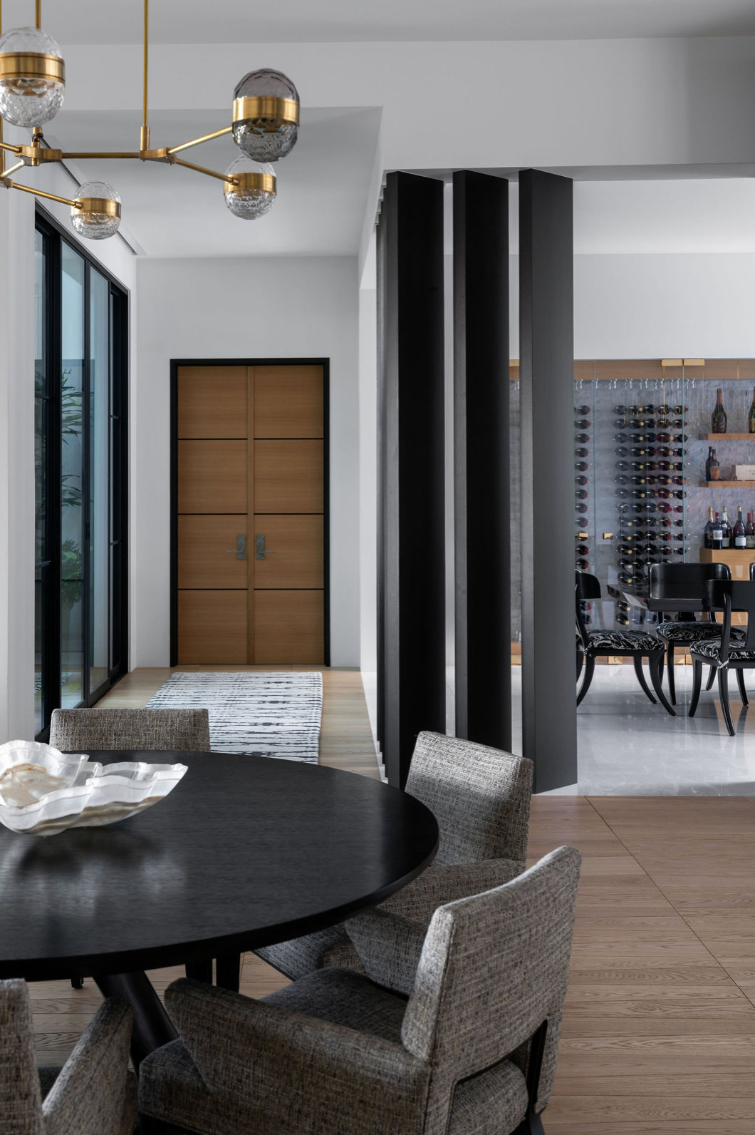 Modern dining area with black round table, gray woven chairs, brass chandelier, and view of wine cellar beyond dark columns.