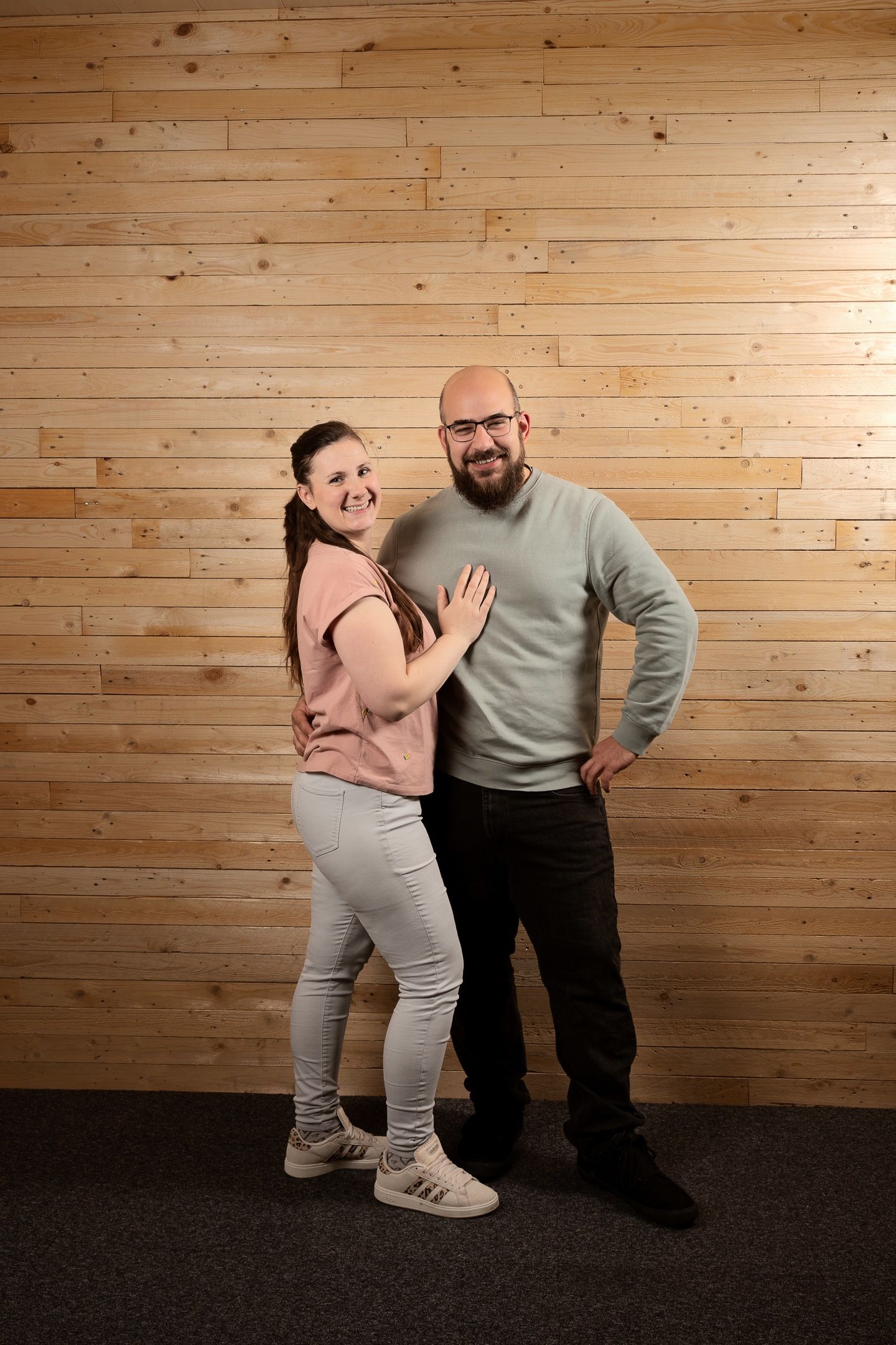 A smiling bald man with glasses and a woman with a ponytail pose together against a light wood plank wall.