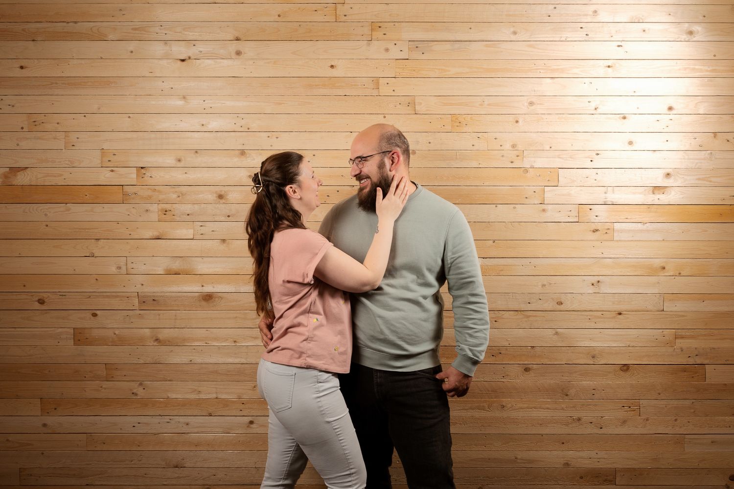 A couple shares a tender moment, smiling at each other against a warm wooden plank wall backdrop.