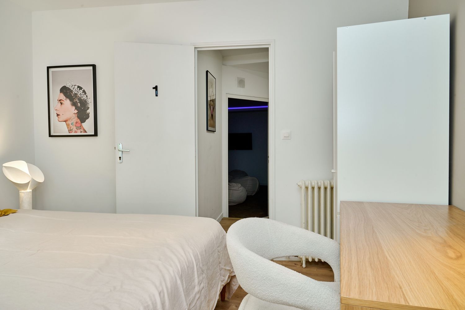 Minimalist white bedroom with a cozy chair, wooden desk, open door revealing hallway, and elegant portrait artwork on the wall.