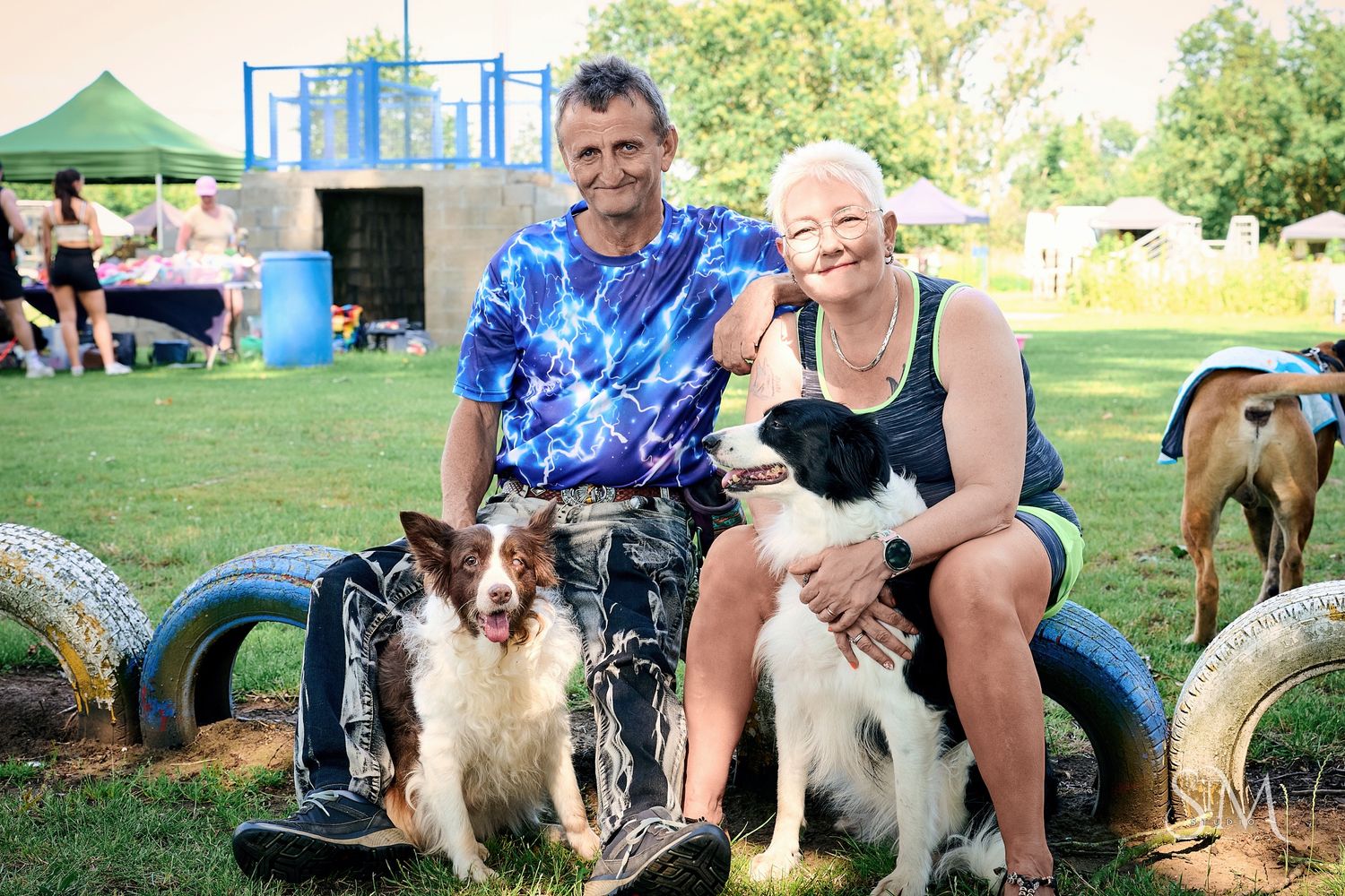 Couple sitting on tires with two border collies at an outdoor dog park on a sunny day.