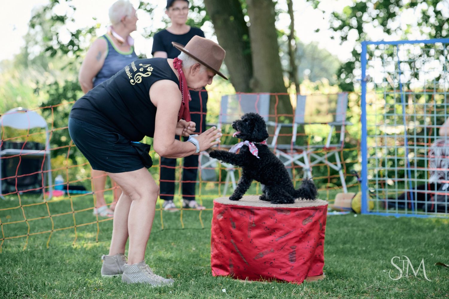 Woman in black shirt and hat training a small black poodle on a red box at an outdoor dog show.