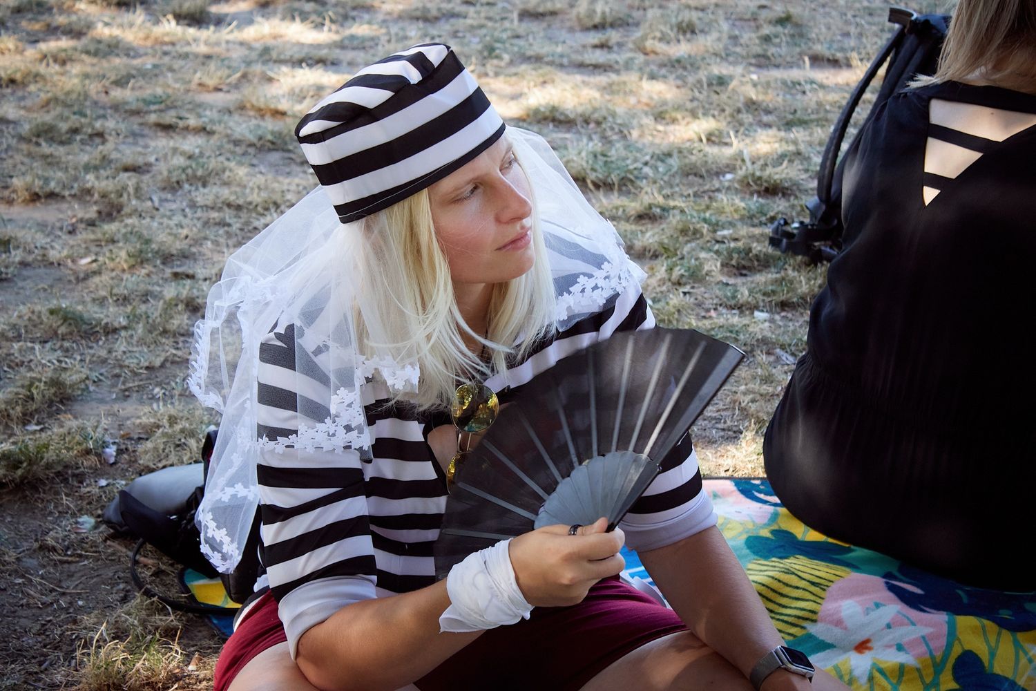 Woman in striped black and white costume with veil hat holds a fan outdoors at a costume event.