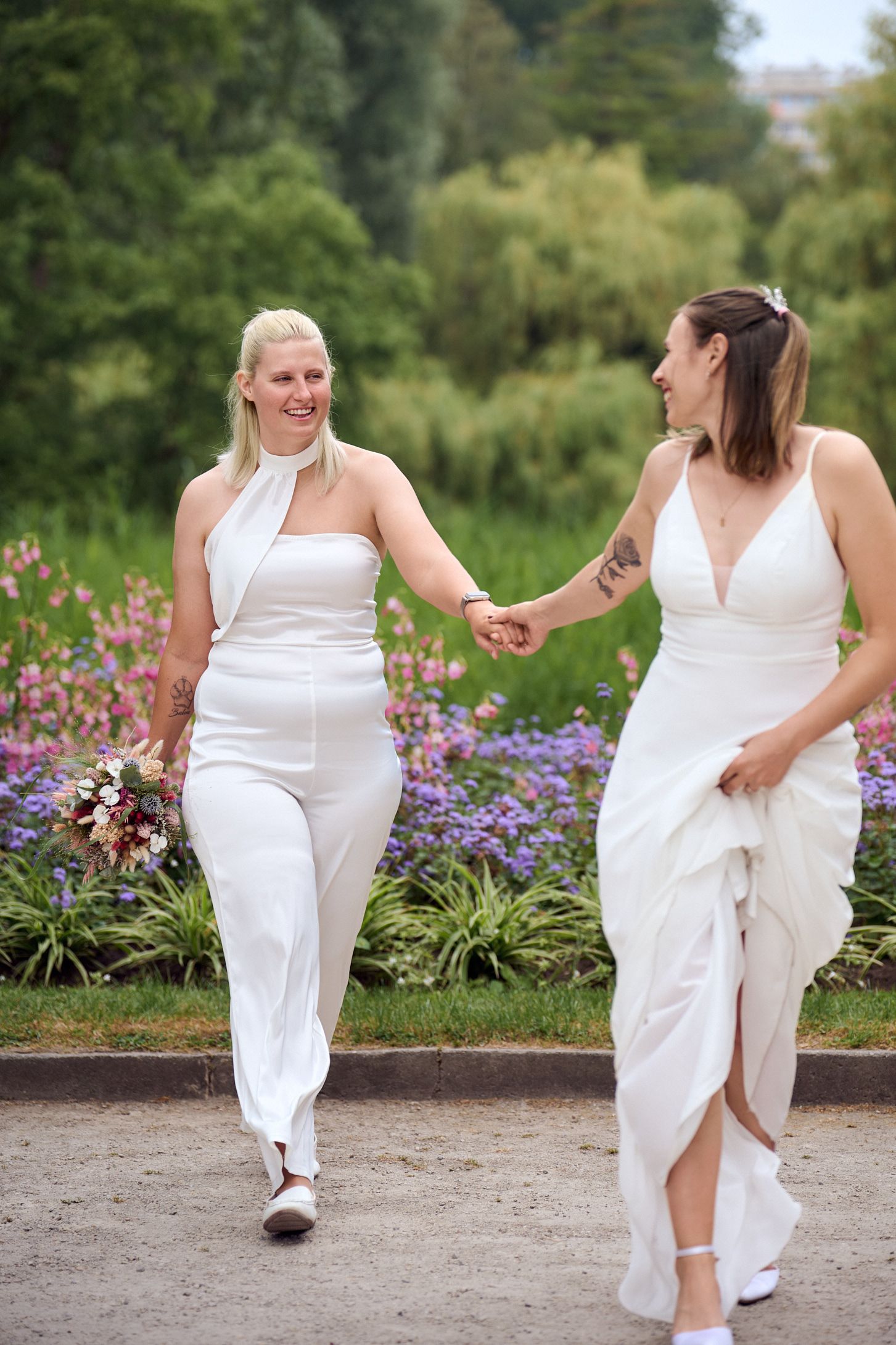 Two brides in white wedding attire hold hands and walk through a flower garden on their wedding day.