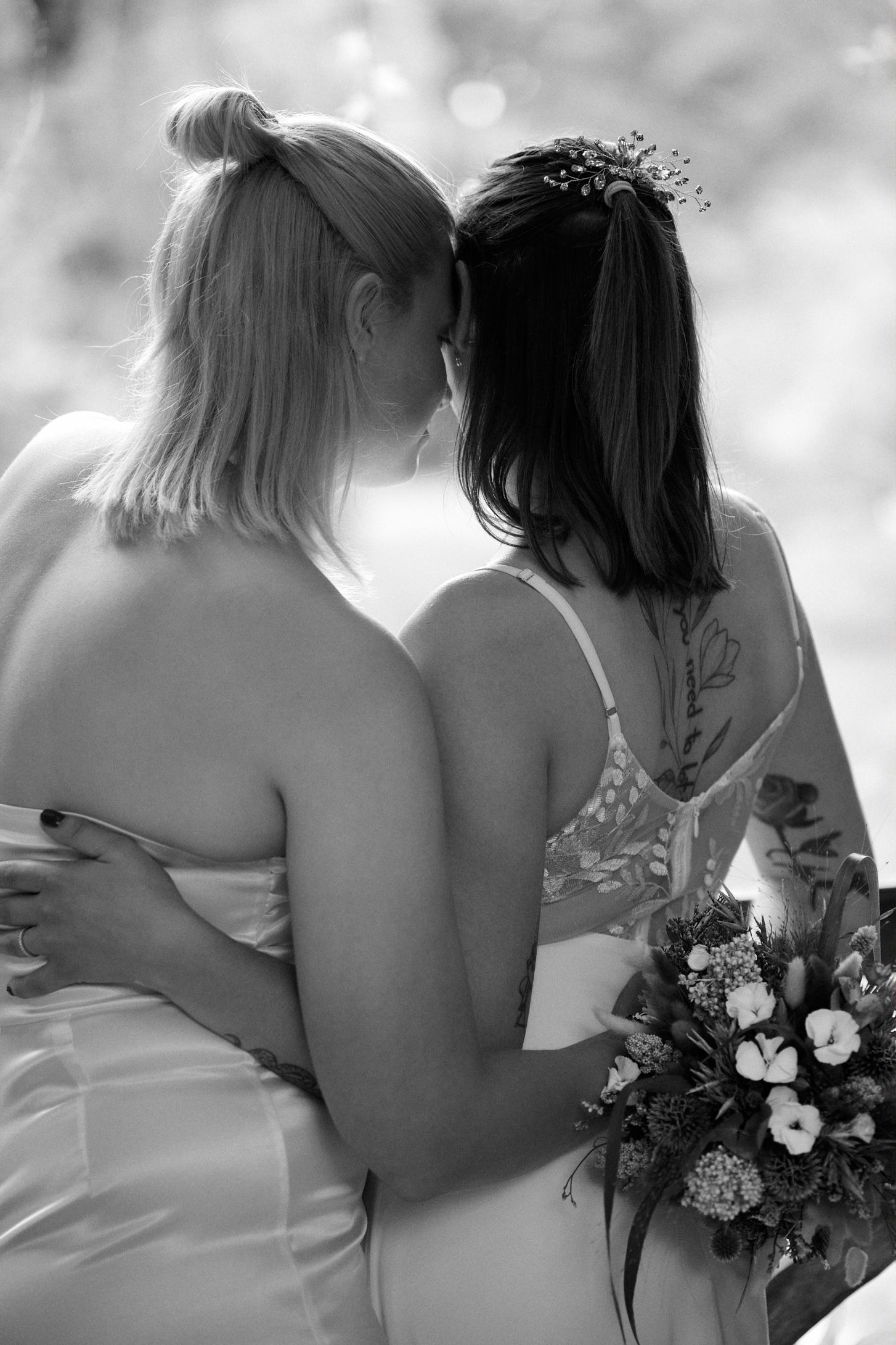 Black and white photo of two brides in white dresses embracing, one holding a bouquet, touching foreheads tenderly.