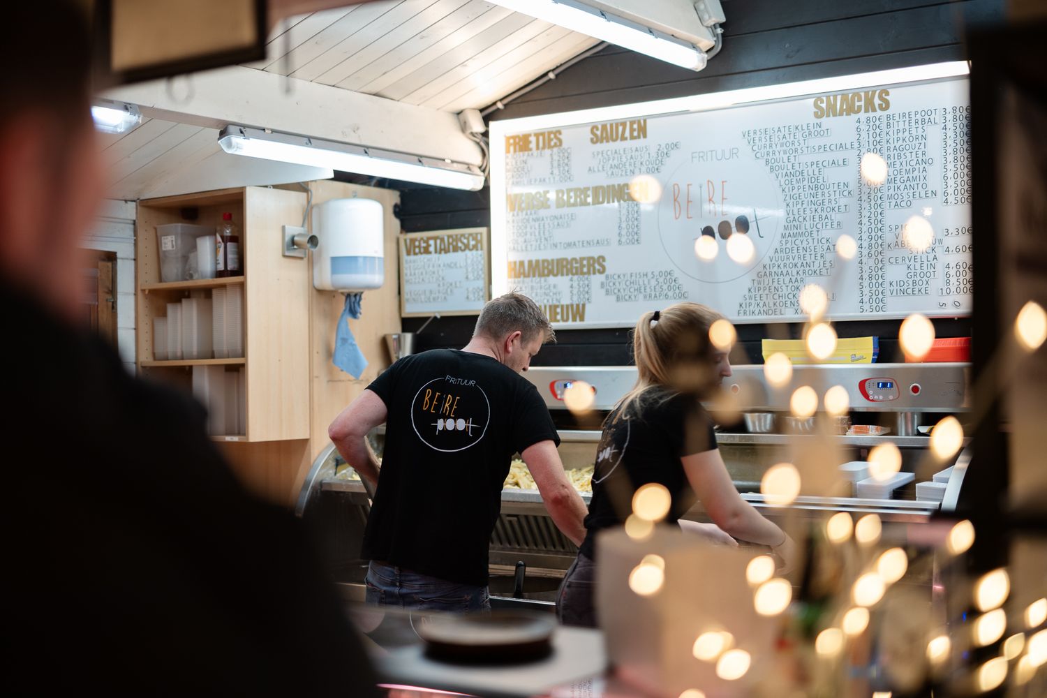 Two staff members working behind a busy restaurant counter with a menu board displayed on the wall above.