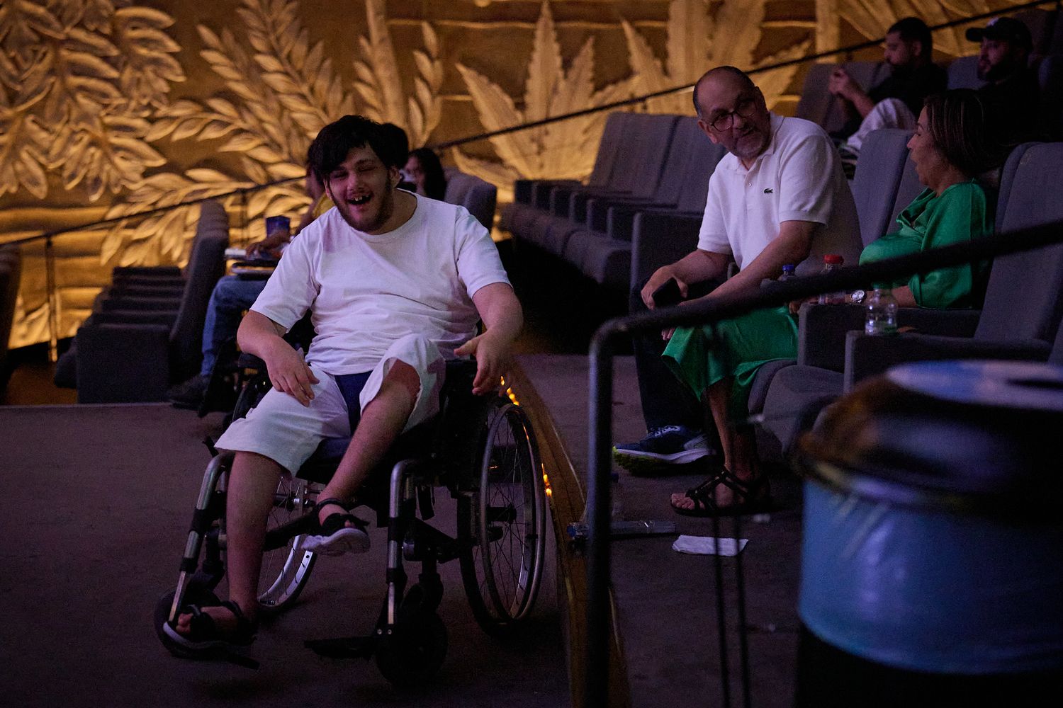 Man in wheelchair inside a dimly lit theater with other audience members seated in rows behind him.