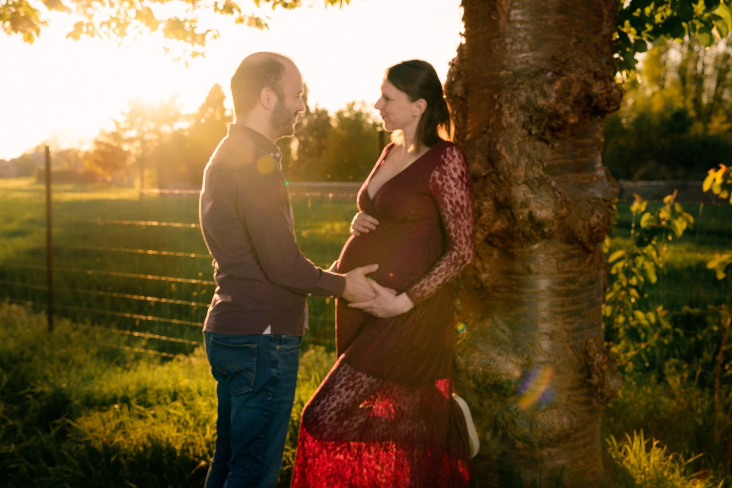 Couple sharing a tender maternity moment by a tree in golden sunset light, man gently touching pregnant woman's belly outdoors.