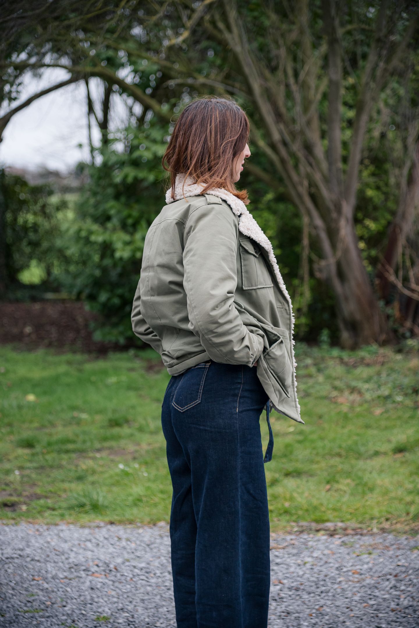 Woman in olive green shearling jacket and dark jeans standing in a garden.