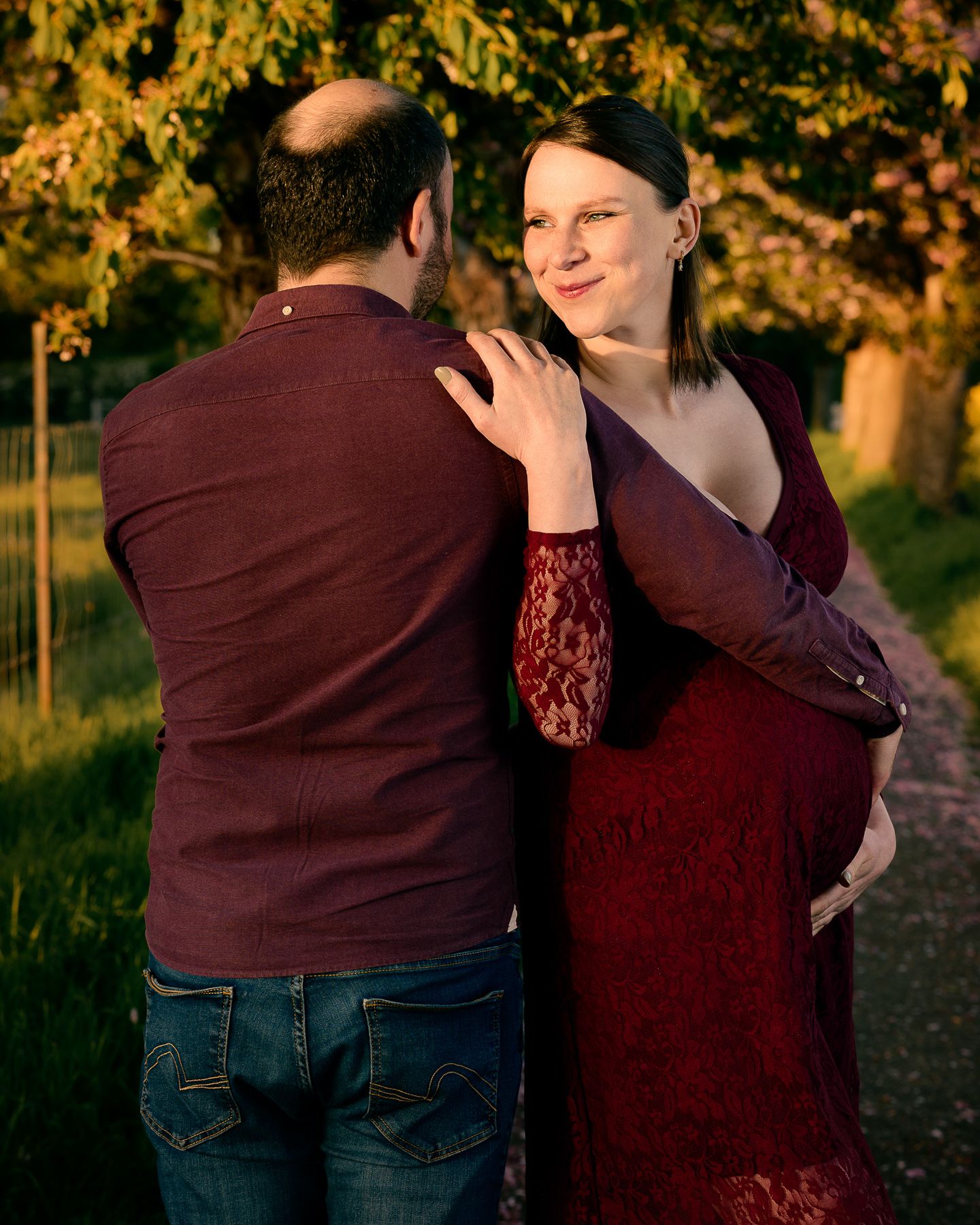 Pregnant woman in red lace dress embracing partner during golden hour maternity photoshoot outdoors.