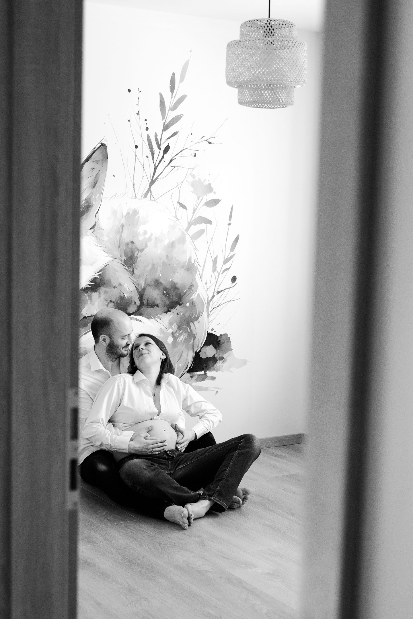 Black and white photo of a couple sitting on floor, man kissing pregnant woman's head against floral wall mural.