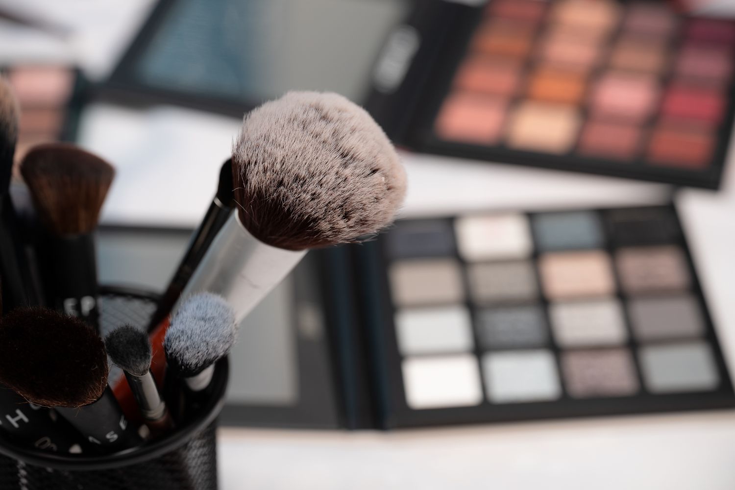 Makeup brushes in a holder with colorful eyeshadow palettes in the background on a white surface.